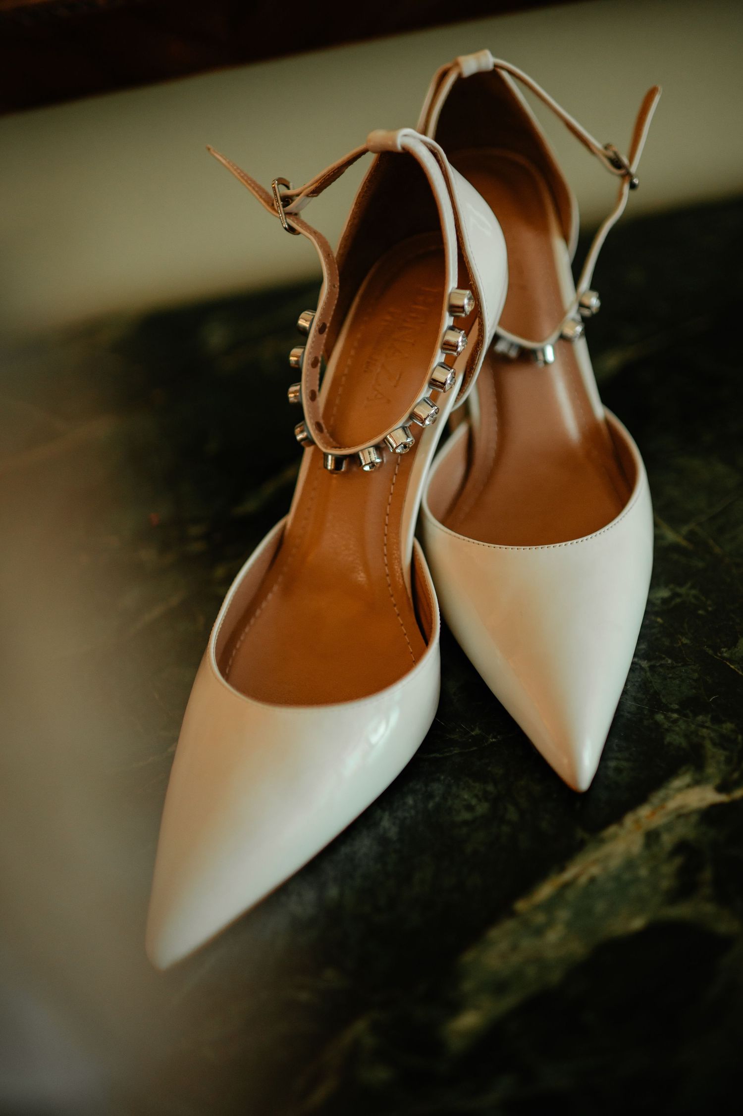 Elegant white pointed-toe bridal heels with crystal ankle strap detail on dark marble surface.