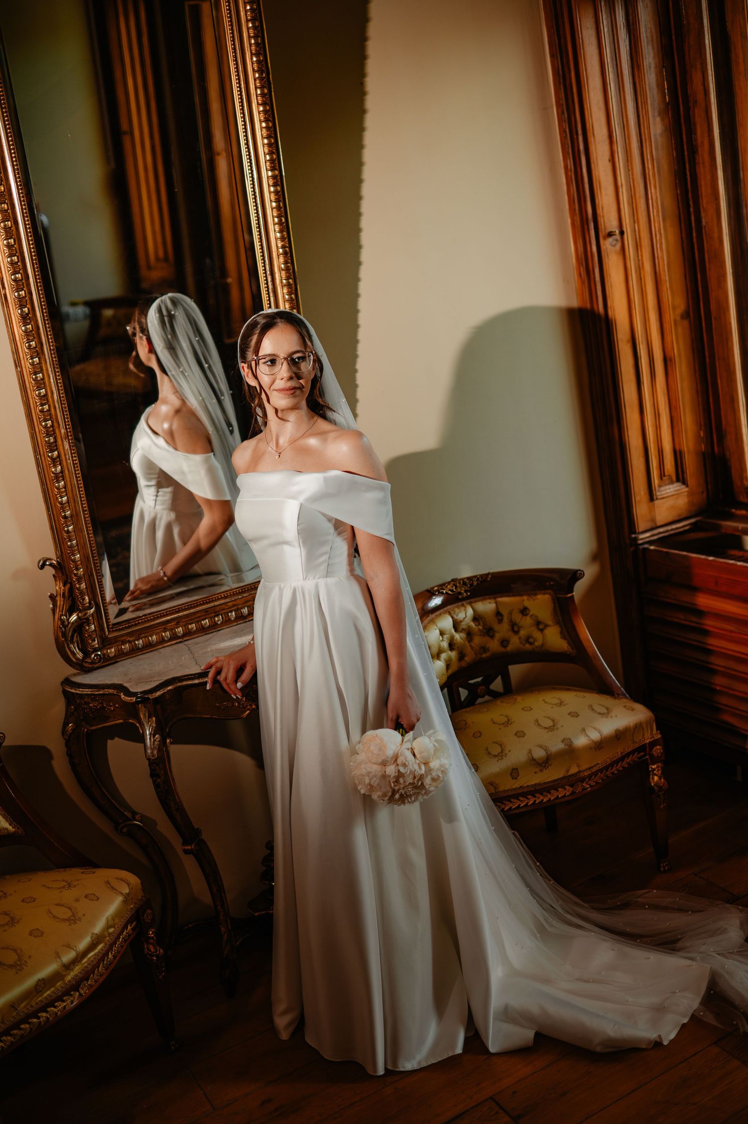 Elegant bride in off-shoulder white satin gown with veil and glasses poses beside ornate gold mirror in vintage room.
