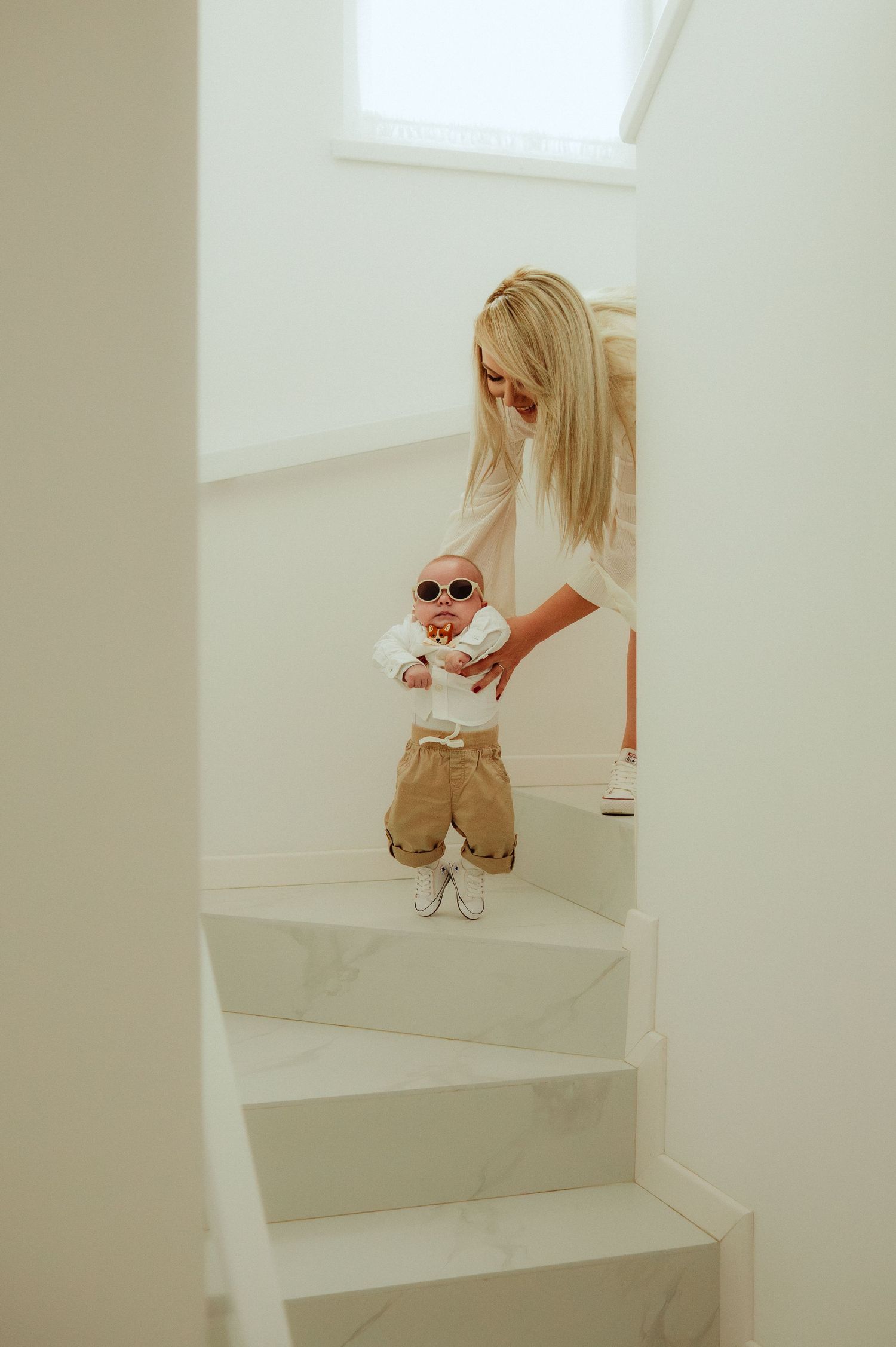 Blonde mother helping stylish baby in sunglasses stand on white marble staircase.
