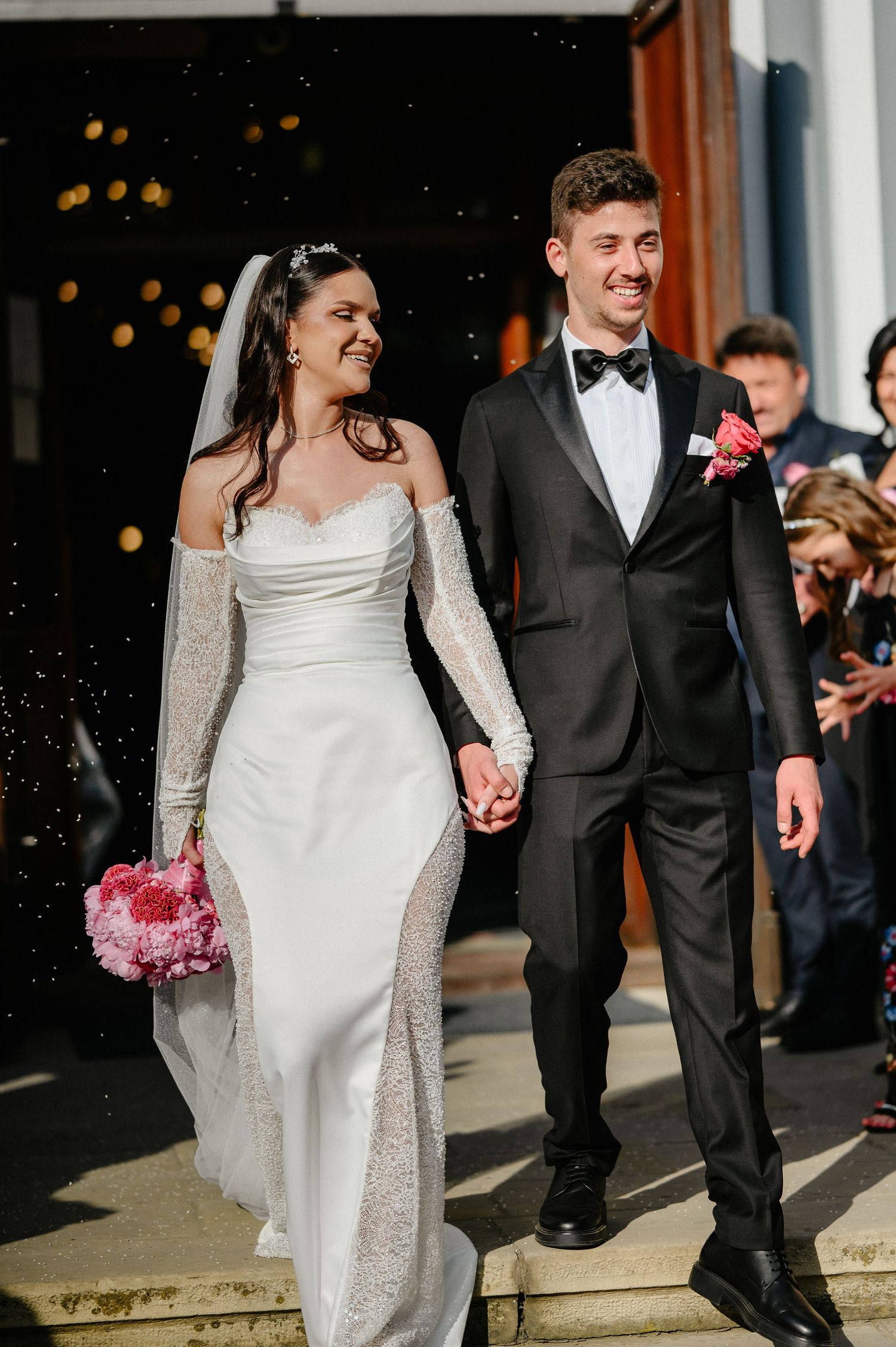 Newlyweds smile and hold hands while descending church steps in bright sunlight after ceremony.