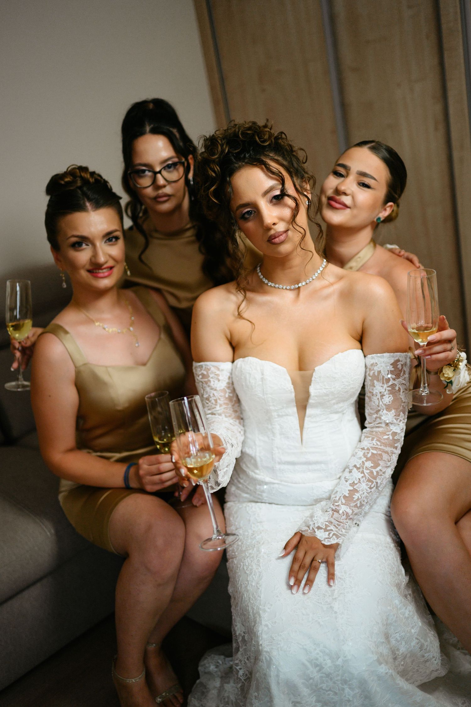 Bride in white lace gown with three bridesmaids in gold dresses, holding champagne glasses, posing together.