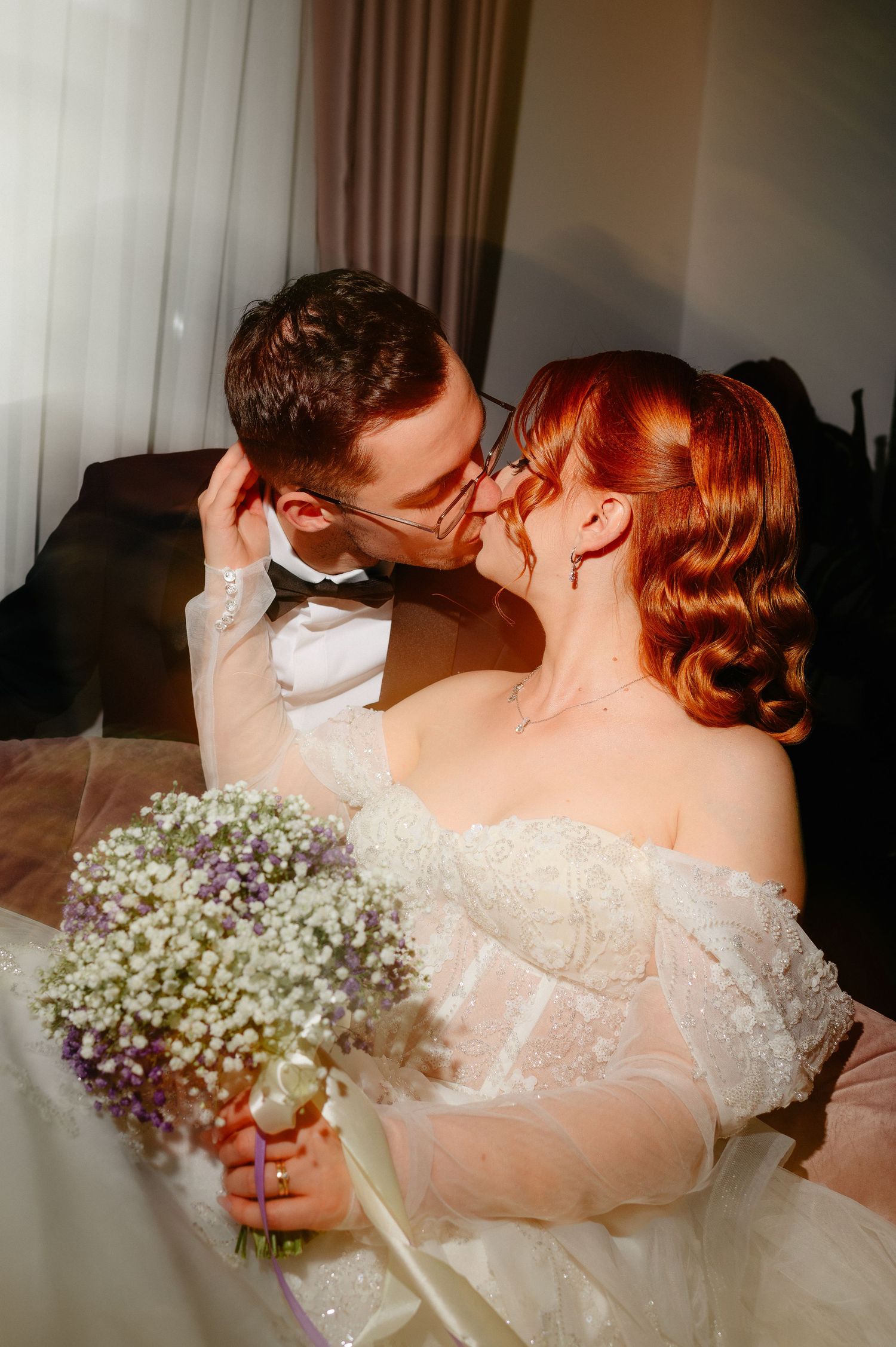 Bride in white lace gown holding baby's breath bouquet shares a kiss with groom in tuxedo in warm sunlit room.