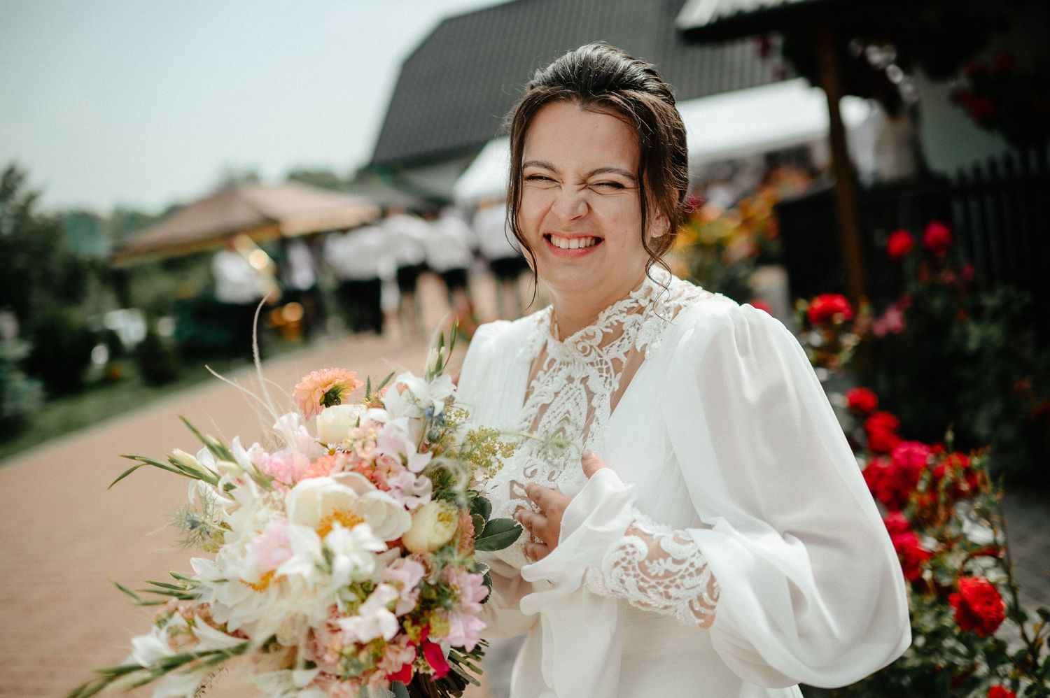 Joyful bride in white lace gown laughs while holding colorful wedding bouquet outdoors.