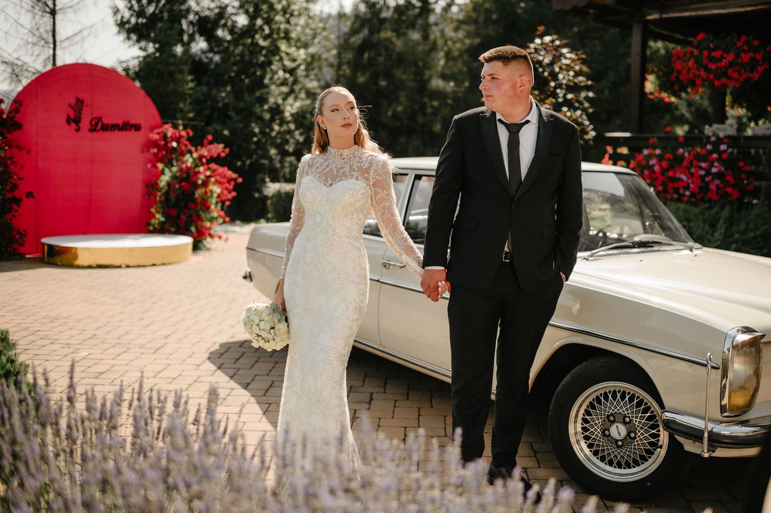 Bride and groom holding hands beside a vintage white Mercedes car at an elegant outdoor wedding venue.