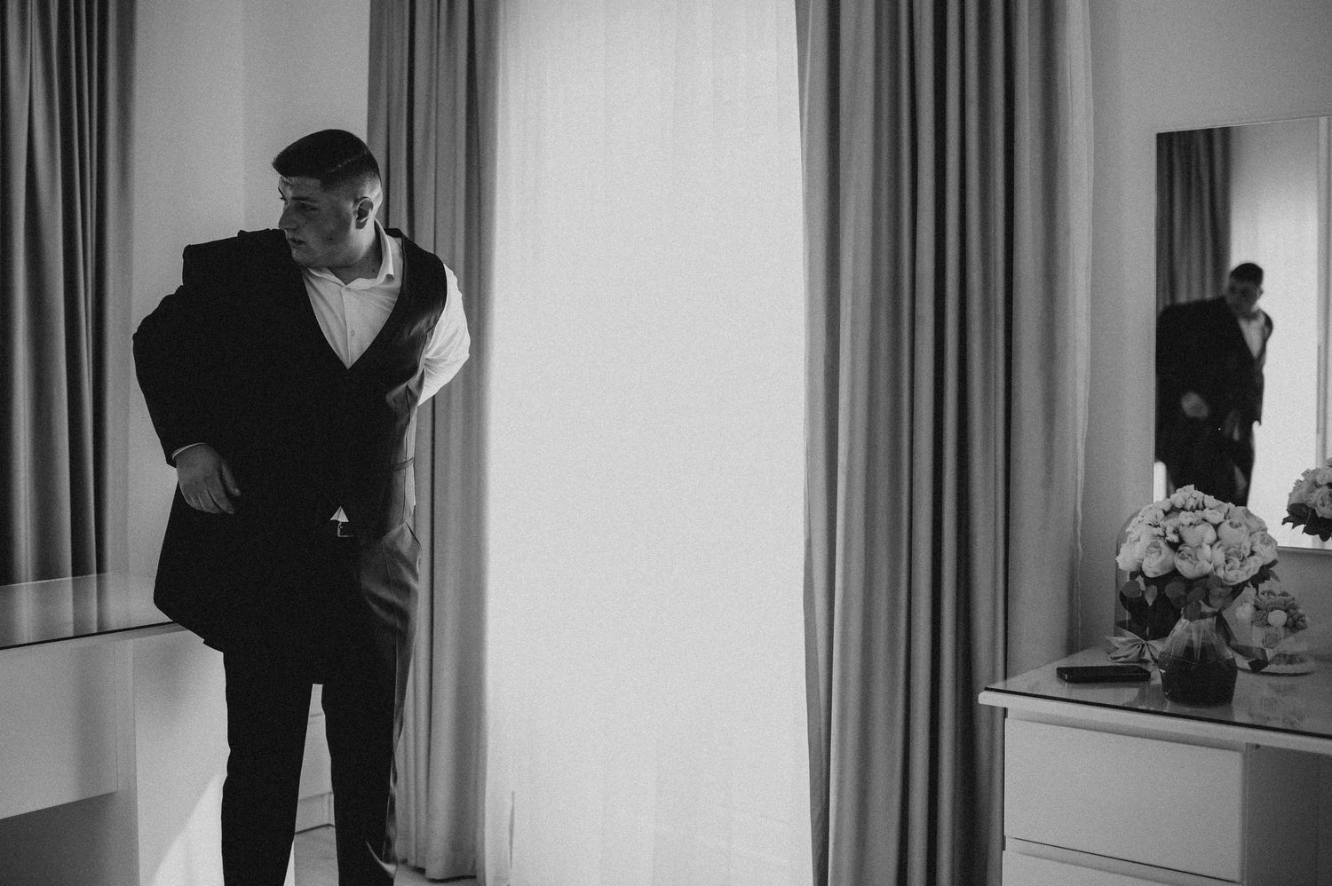 Black and white photo of a man in formal attire adjusting his jacket near a window, with his reflection visible in a mirror.