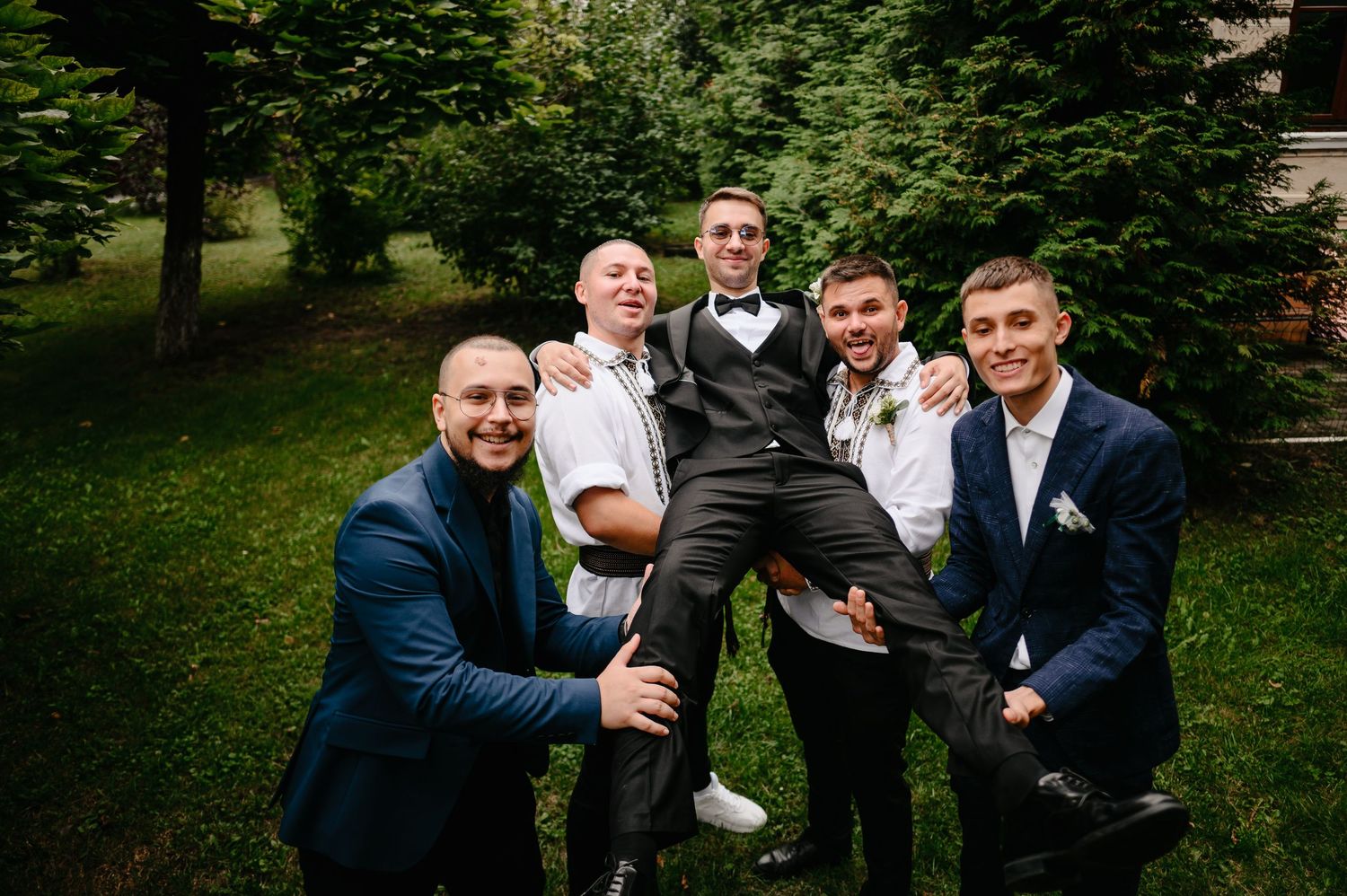 Four groomsmen in navy suits lifting and celebrating the groom in a black tuxedo outdoors at a wedding.