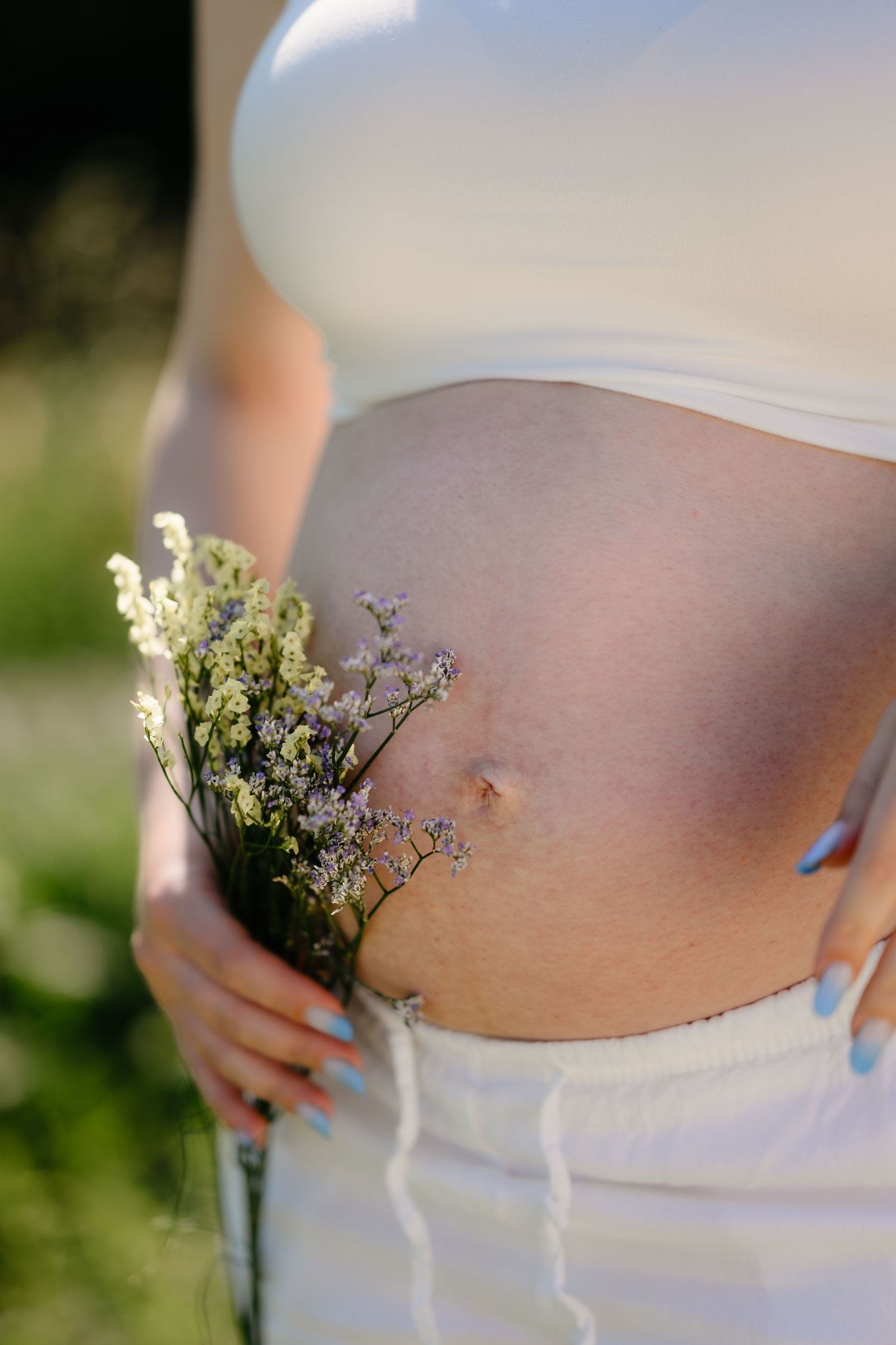 Close-up of pregnant belly with delicate wildflowers tucked into white pants waistband outdoors.