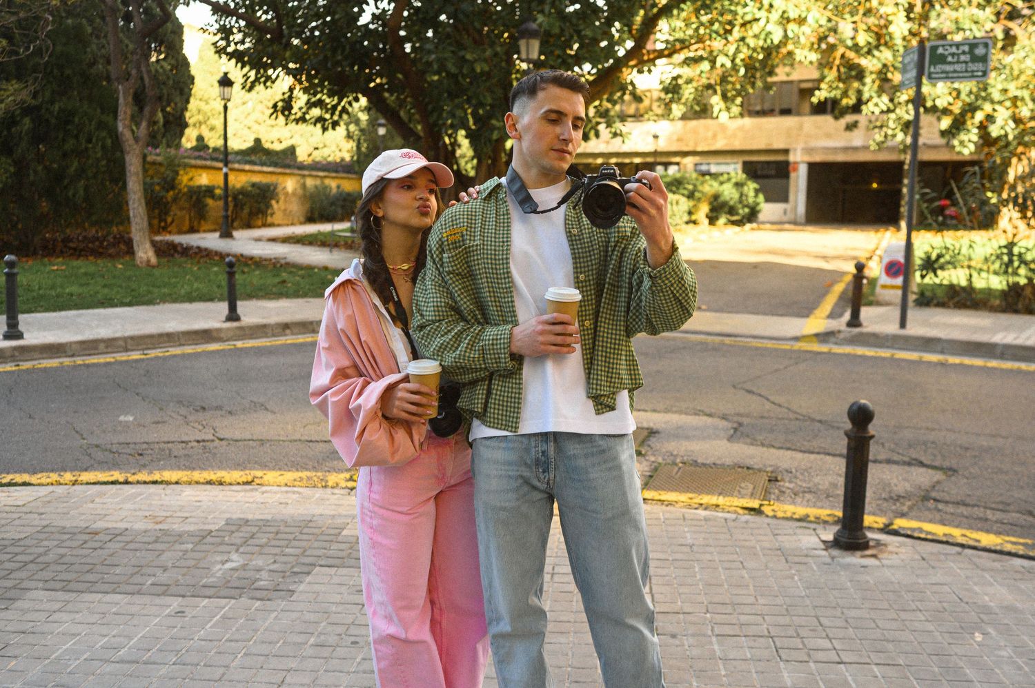 Young couple posing on a street, woman in pink outfit holding coffee, man in green jacket holding a camera.