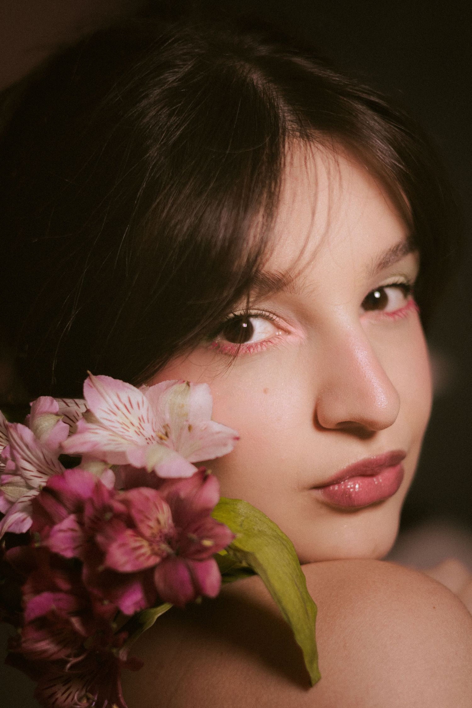 Close-up portrait of a young woman with pink eye makeup resting her face near pink flowers.