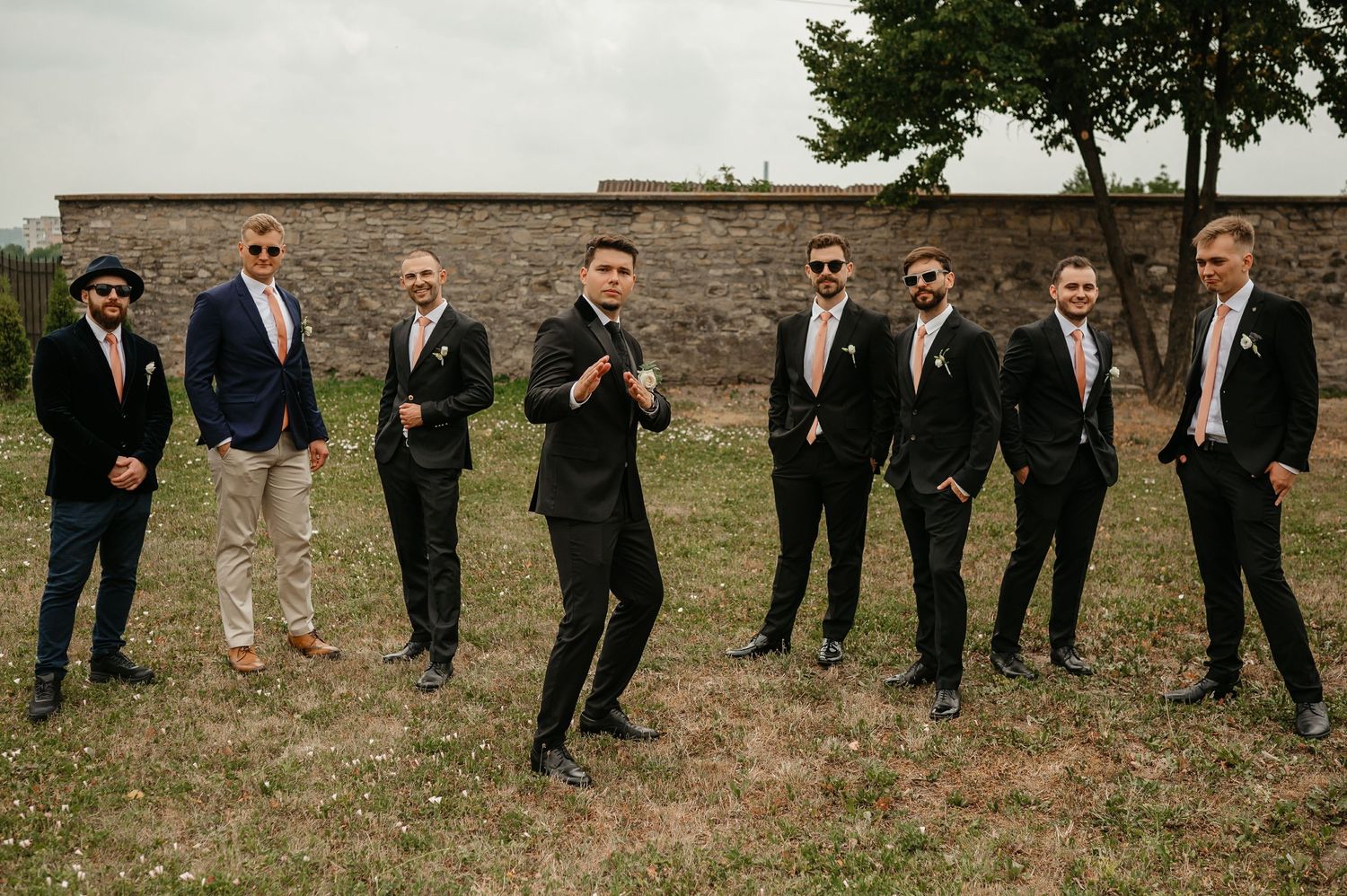 Groomsmen in suits and ties posing outdoors on grass with a stone wall backdrop at a wedding.