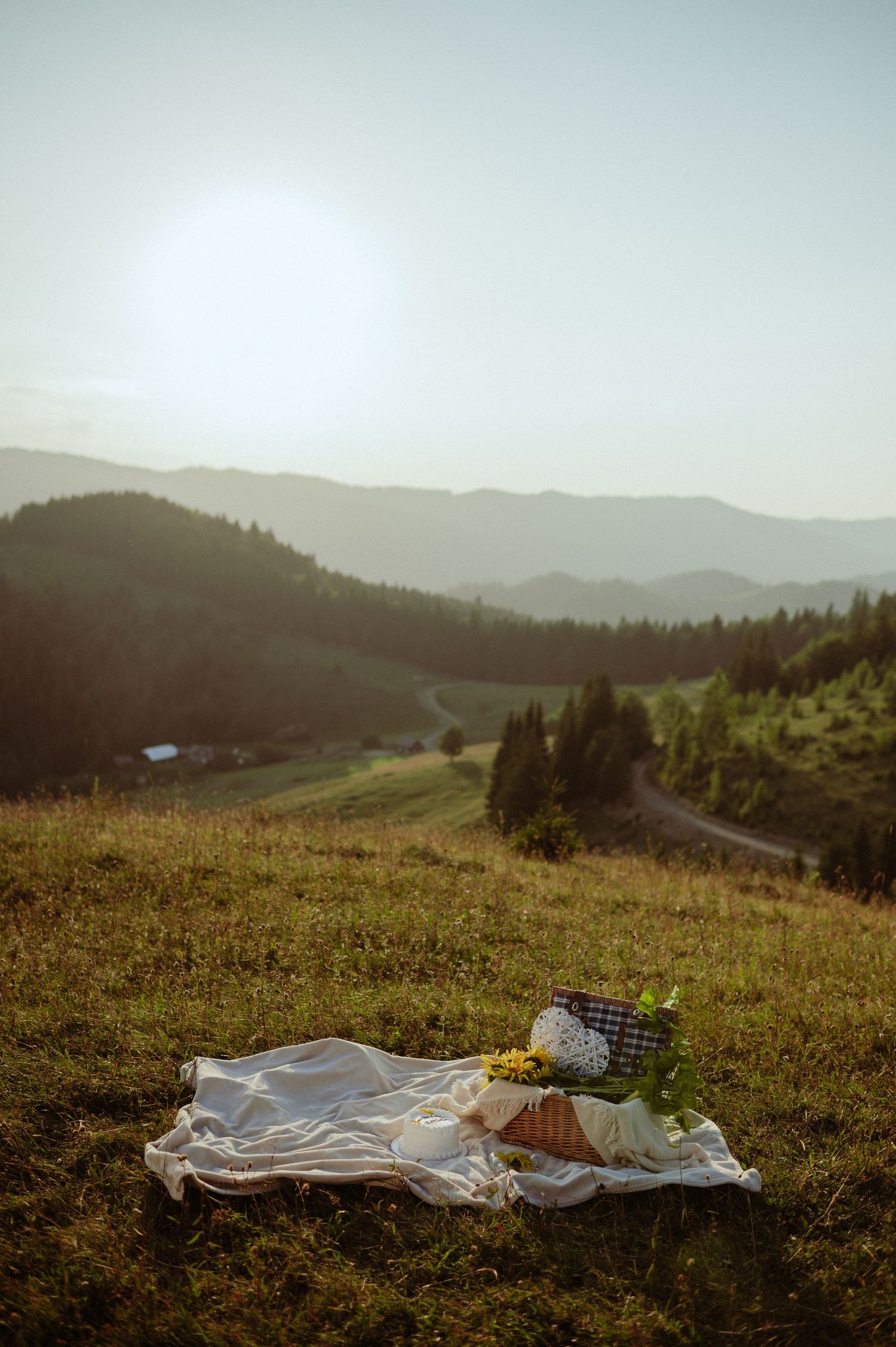 Romantic picnic setup on green hillside with mountain valley sunset backdrop.