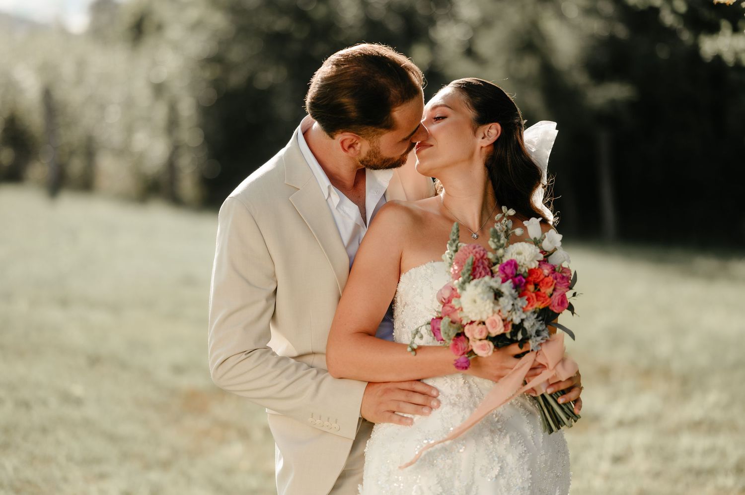 Groom kissing bride on the cheek outdoors as she smiles holding a colorful bridal bouquet.
