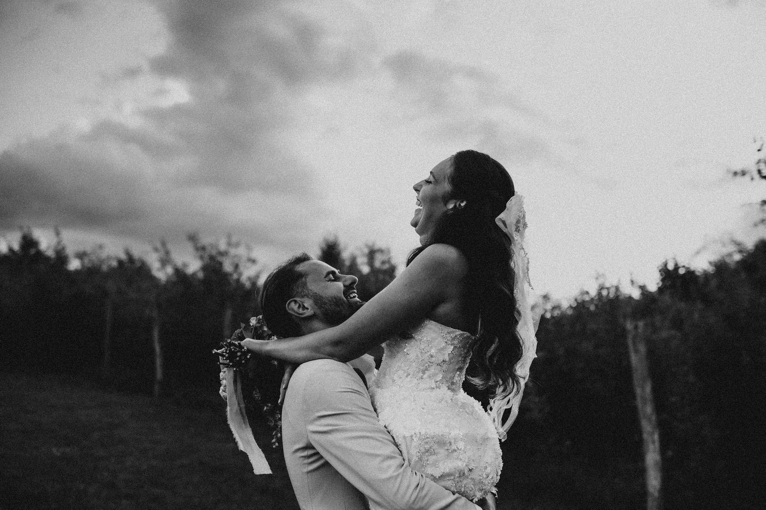 Black and white photo of groom lifting bride outdoors under cloudy dramatic sky.