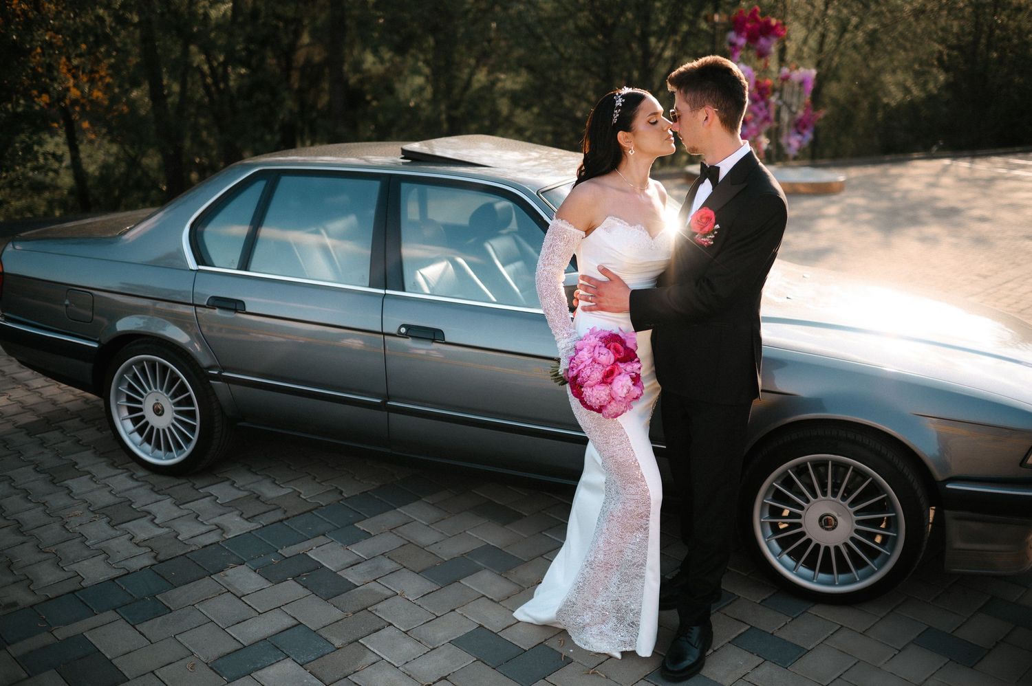 Bride and groom embracing beside classic gray BMW with pink peony bouquet outdoors.
