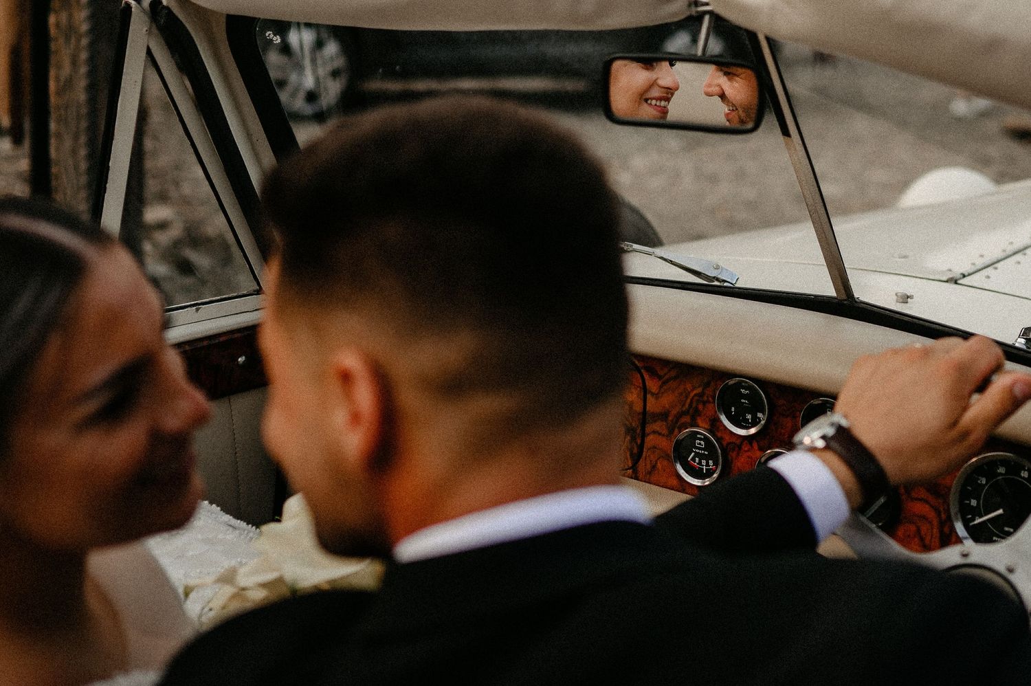 Romantic reflection of bride and groom kissing captured in vintage car rearview mirror.