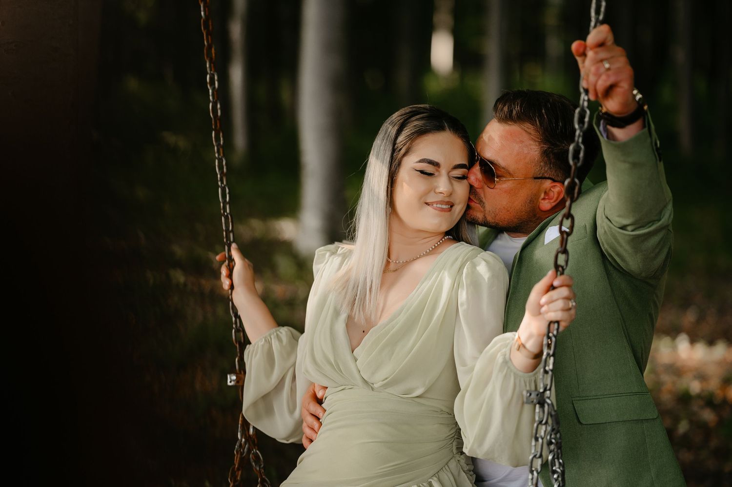 Couple sharing a romantic moment on a chain swing in a forest, man in green blazer kissing her cheek.