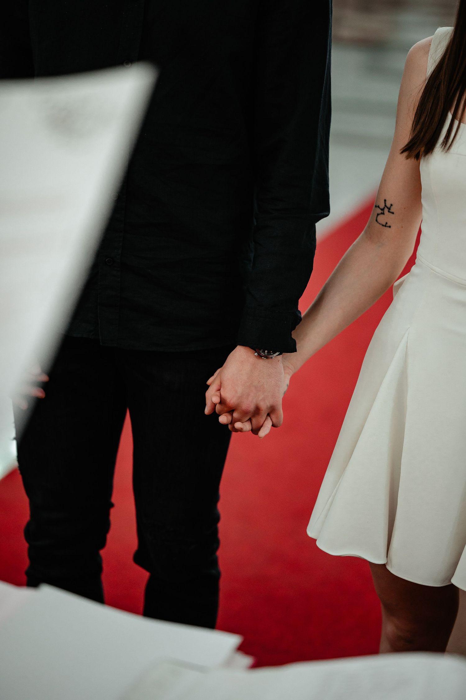 Close-up of newlyweds holding hands, bride in white dress with anchor tattoo, groom in black, standing on red carpet.