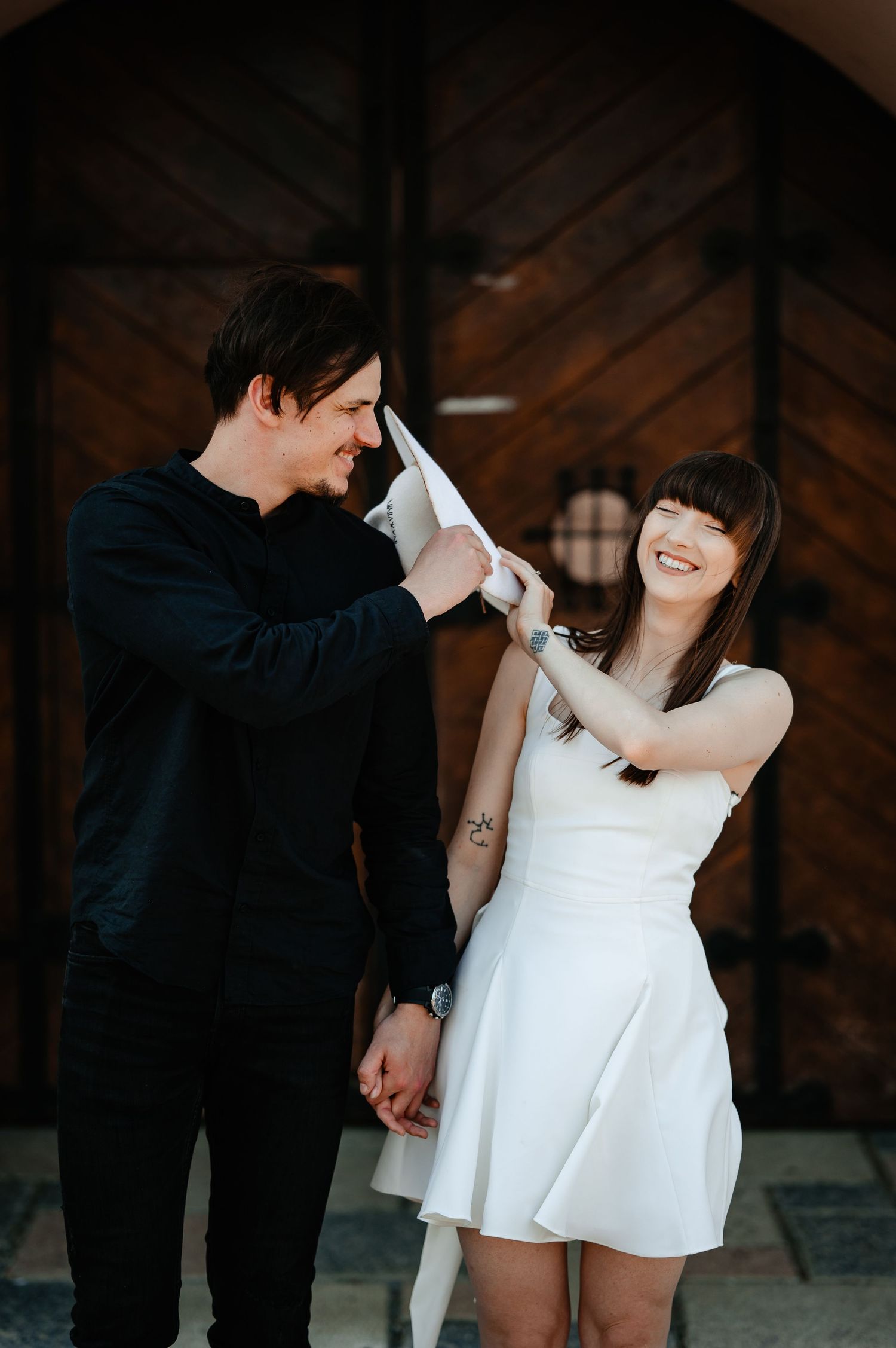 Laughing couple playfully sharing a white hat while holding hands before a wooden door.