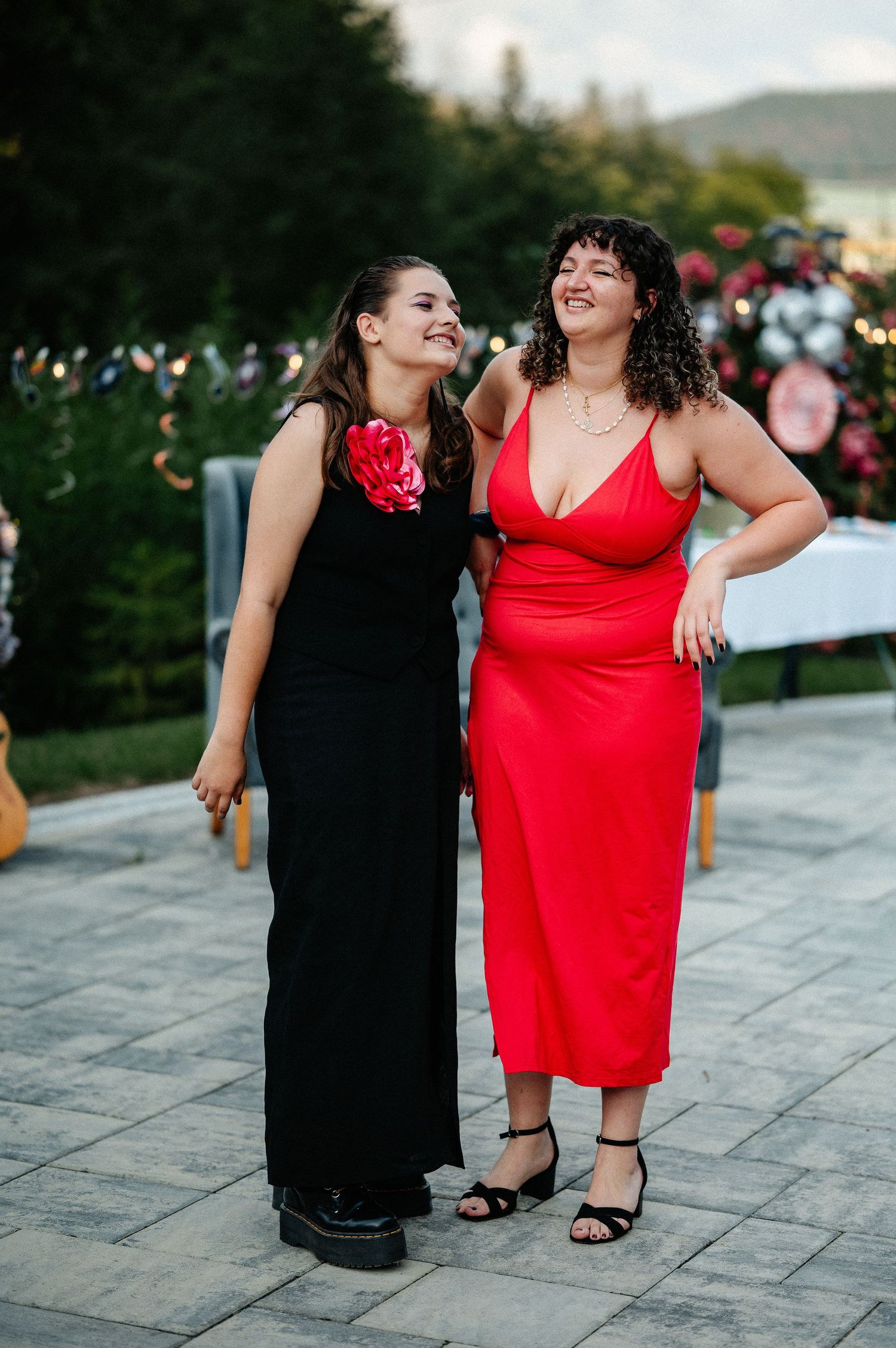 Two women smiling together outdoors, one in a black gown and one in a red midi dress at a party.