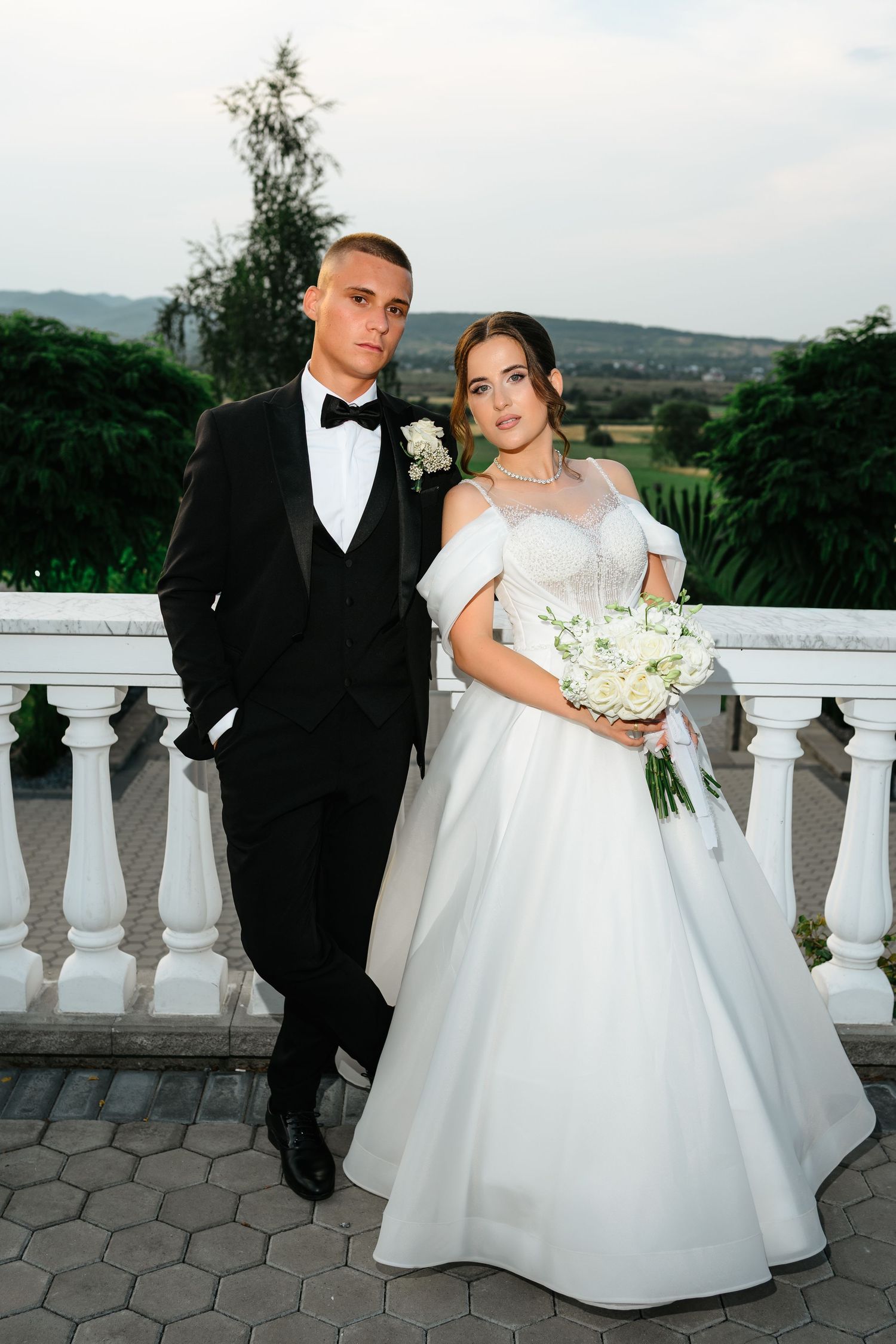 Elegant bride and groom posing on a terrace, bride in white ball gown holding bouquet, groom in black tuxedo.