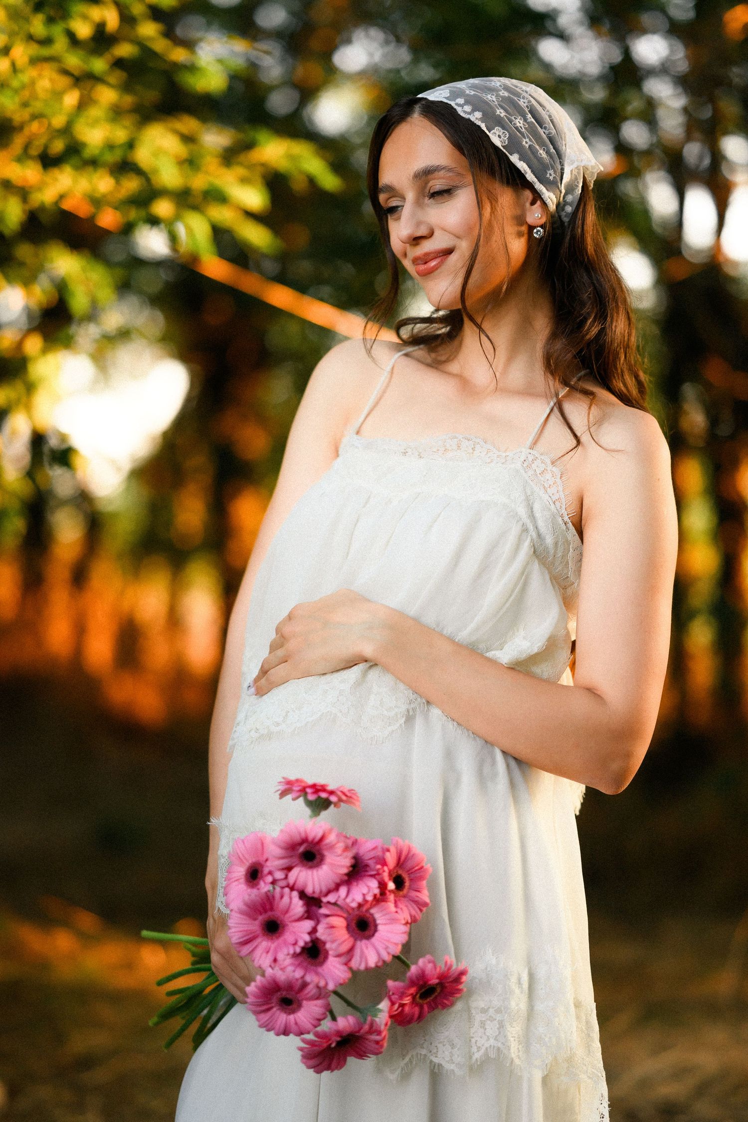 Pregnant woman in white lace dress holding pink gerbera daisies, smiling outdoors in golden sunlight.