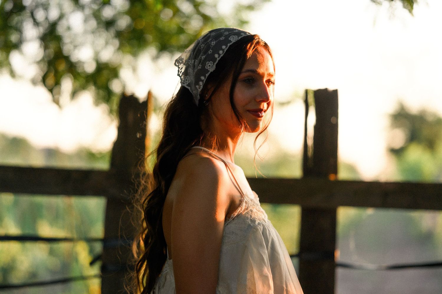 Young woman in white dress and bandana smiles softly at sunset near a rustic wooden fence.