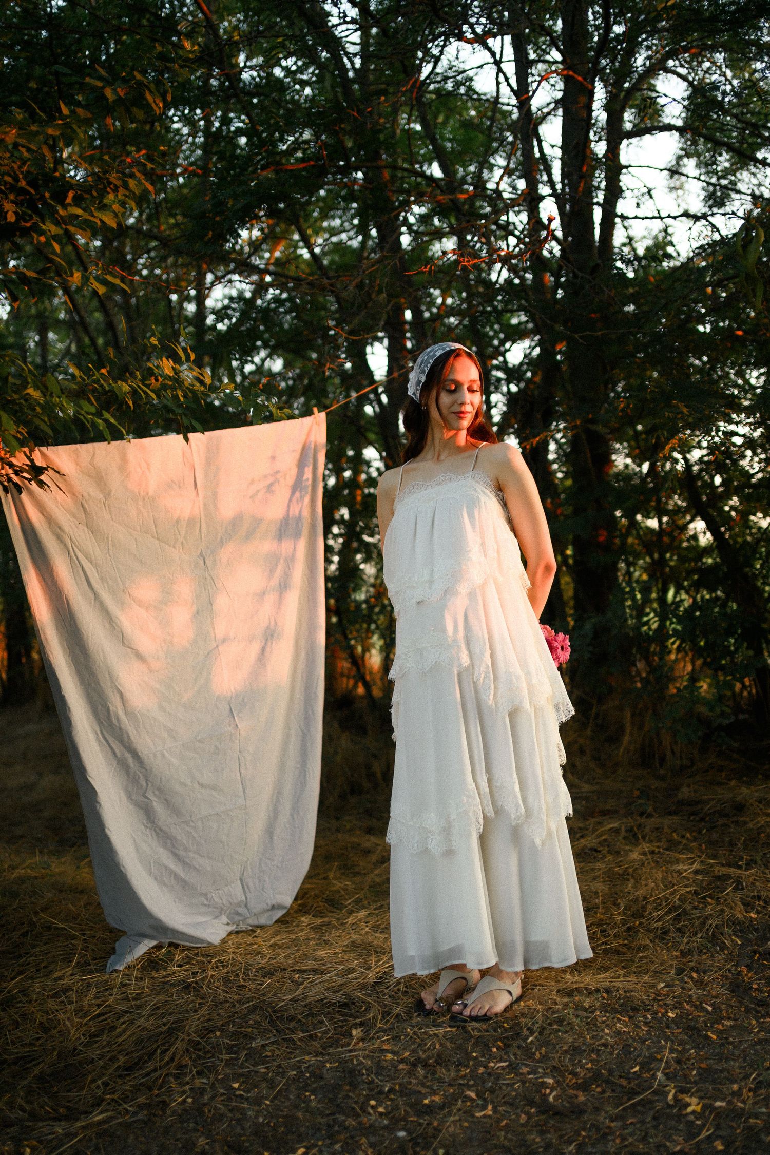 Woman in white tiered dress and headband stands near hanging fabric among trees in golden hour light.