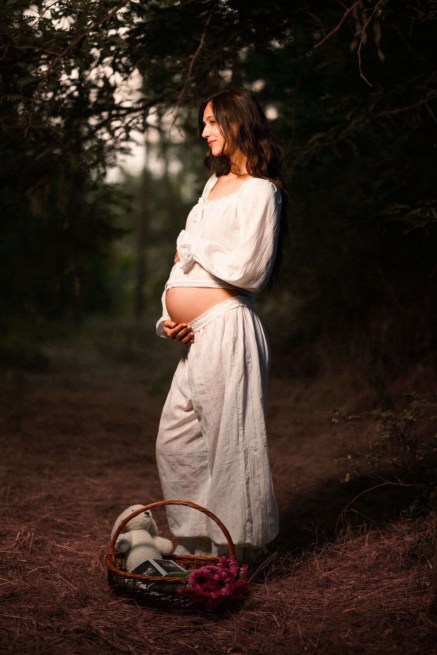 Pregnant woman in white dress cradling baby bump in forest with flower basket, glowing in soft sunlight.