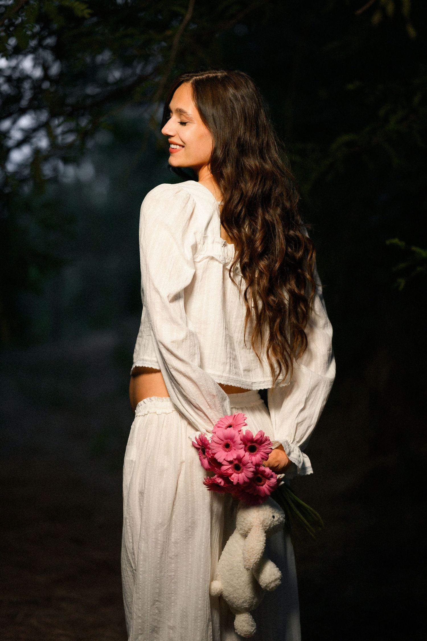 Young woman in white dress holds pink gerbera flowers and stuffed bunny, smiling in a dark forest setting.