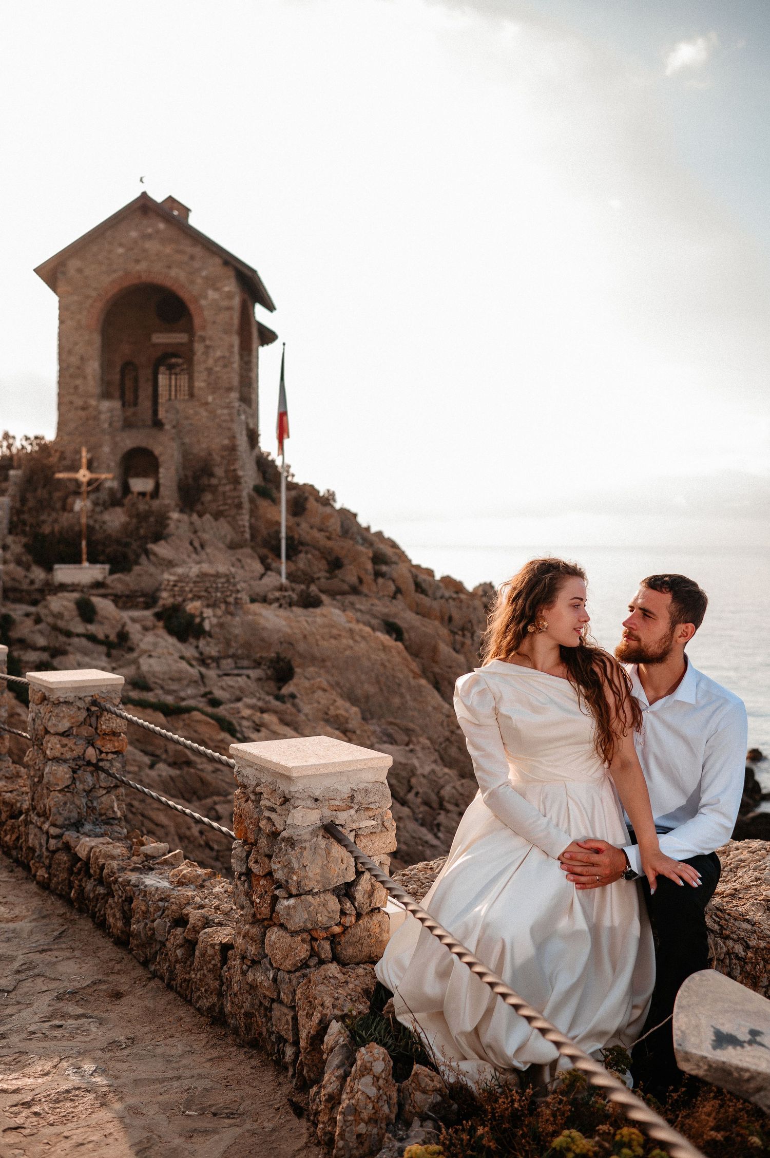 Wedding couple sitting by stone chapel on rocky Italian coastal promontory at sunset.