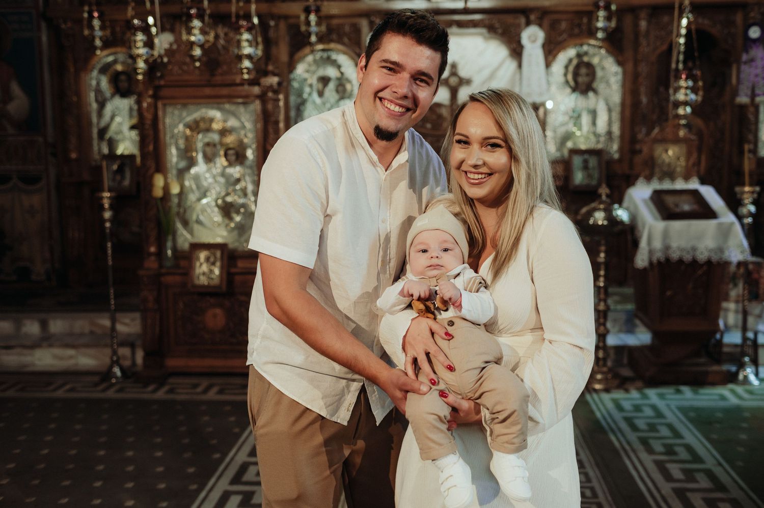 Young family poses with baby in ornate Orthodox church during christening ceremony.