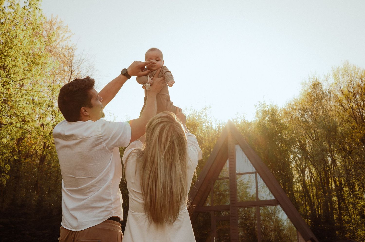 Young couple lifting their baby in the air outdoors in golden sunlight near a modern A-frame house.