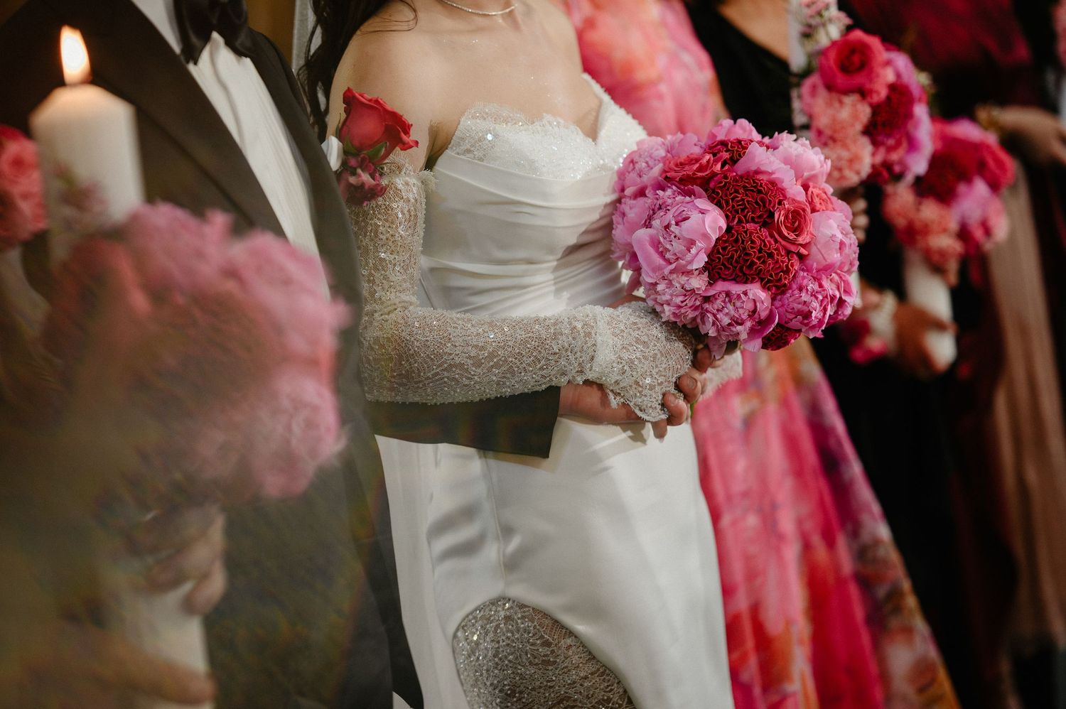 Bride in white gown with lace gloves holds pink peony bouquet beside groom at wedding ceremony.