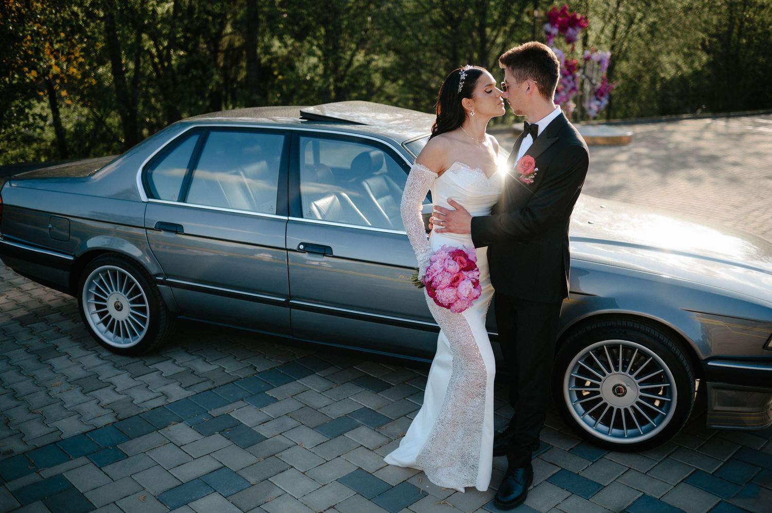 Wedding couple sharing romantic moment beside vintage gray BMW sedan on cobblestone driveway.