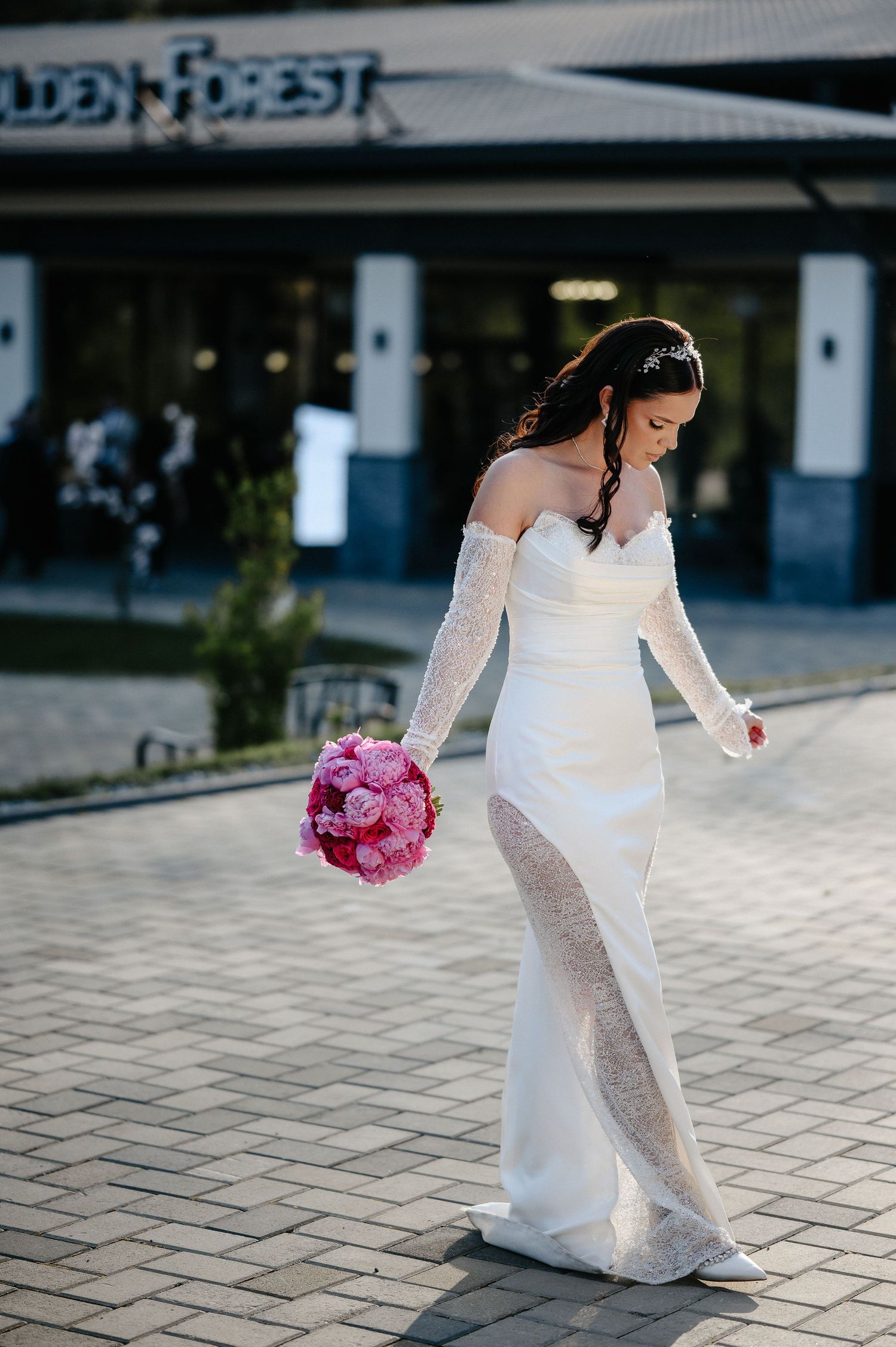 Elegant bride in white lace off-shoulder wedding gown walking alone outside Golden Forest, holding pink bouquet.