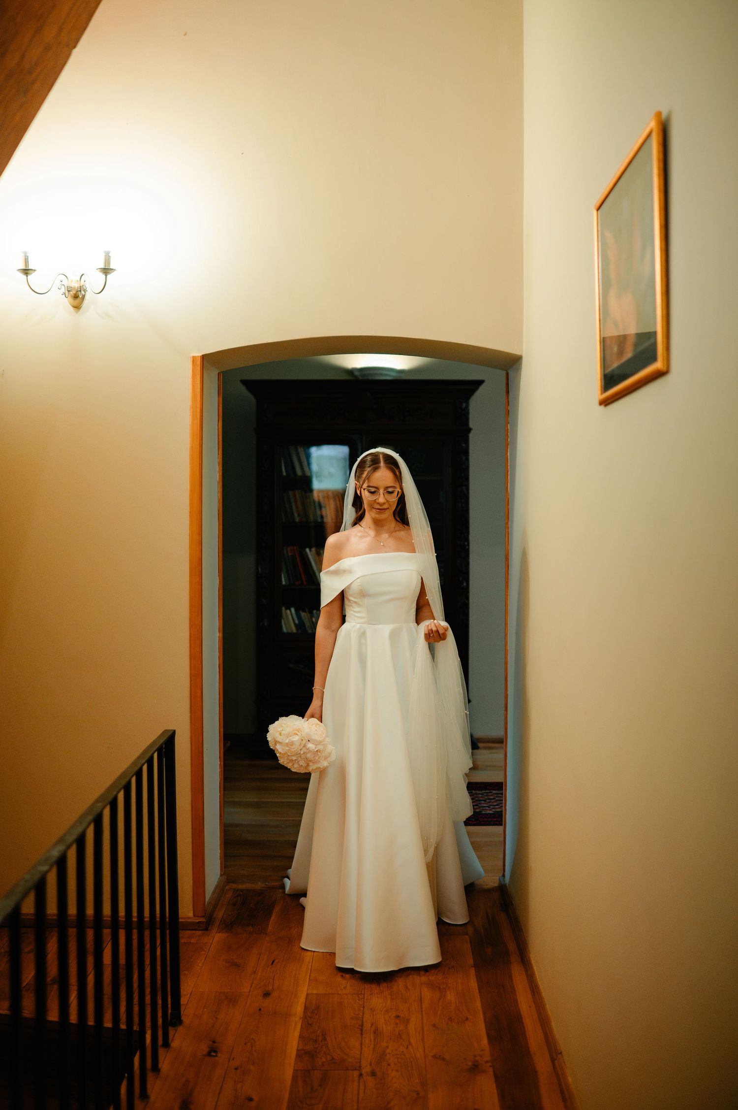 Bride in off-shoulder white wedding dress and veil stands in arched doorway of elegant home.