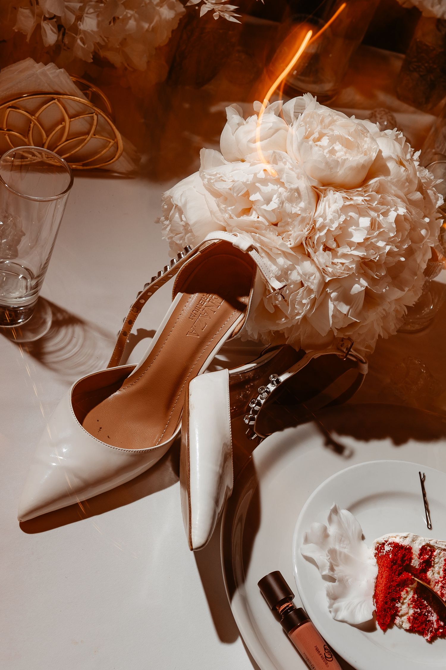 White bridal heels with crystal anklet beside a lush peony bouquet and red velvet cake slice on reception table.