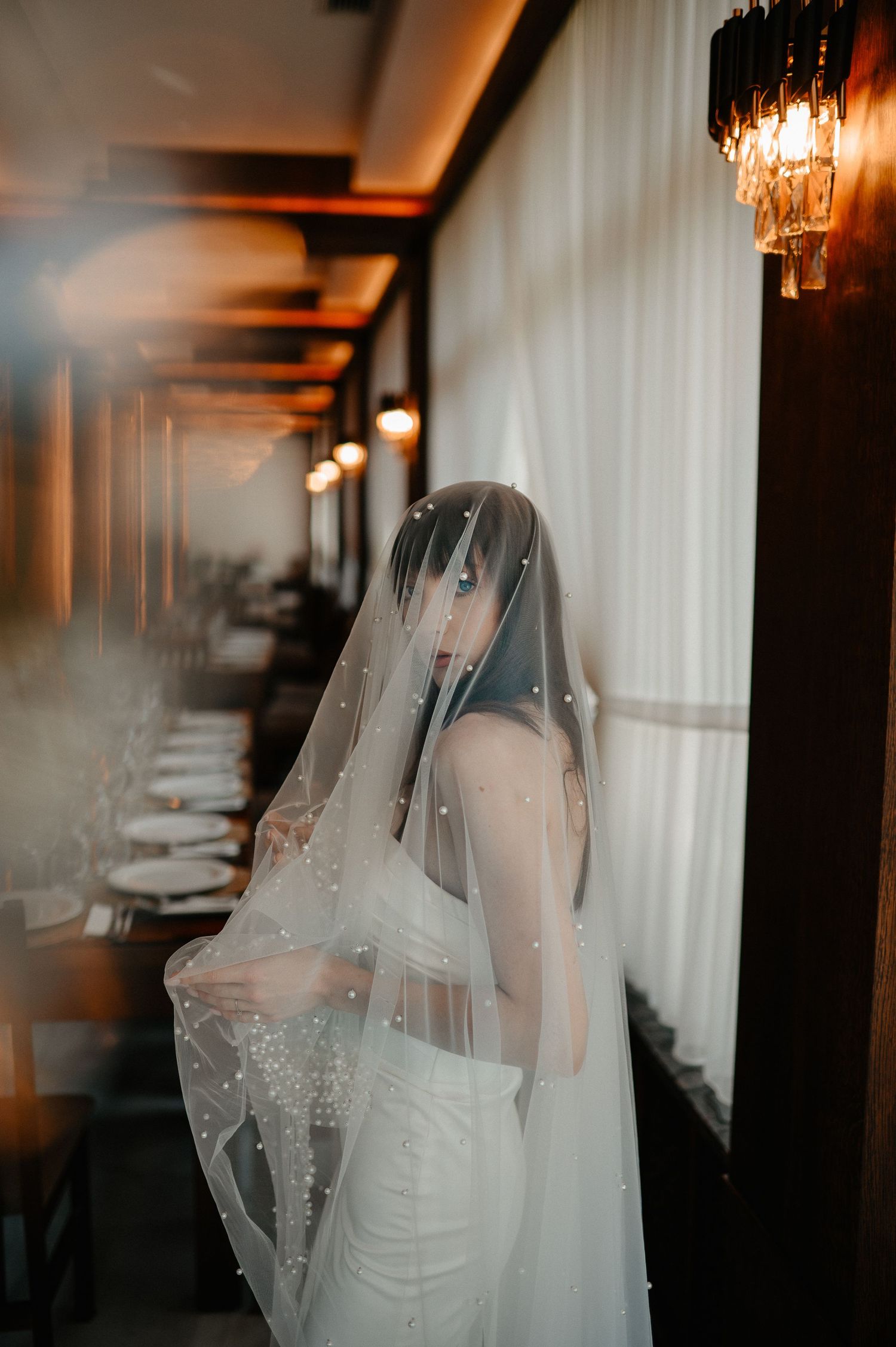 Bride draped in pearl-trimmed veil stands in elegant restaurant corridor with warm ambient lighting.