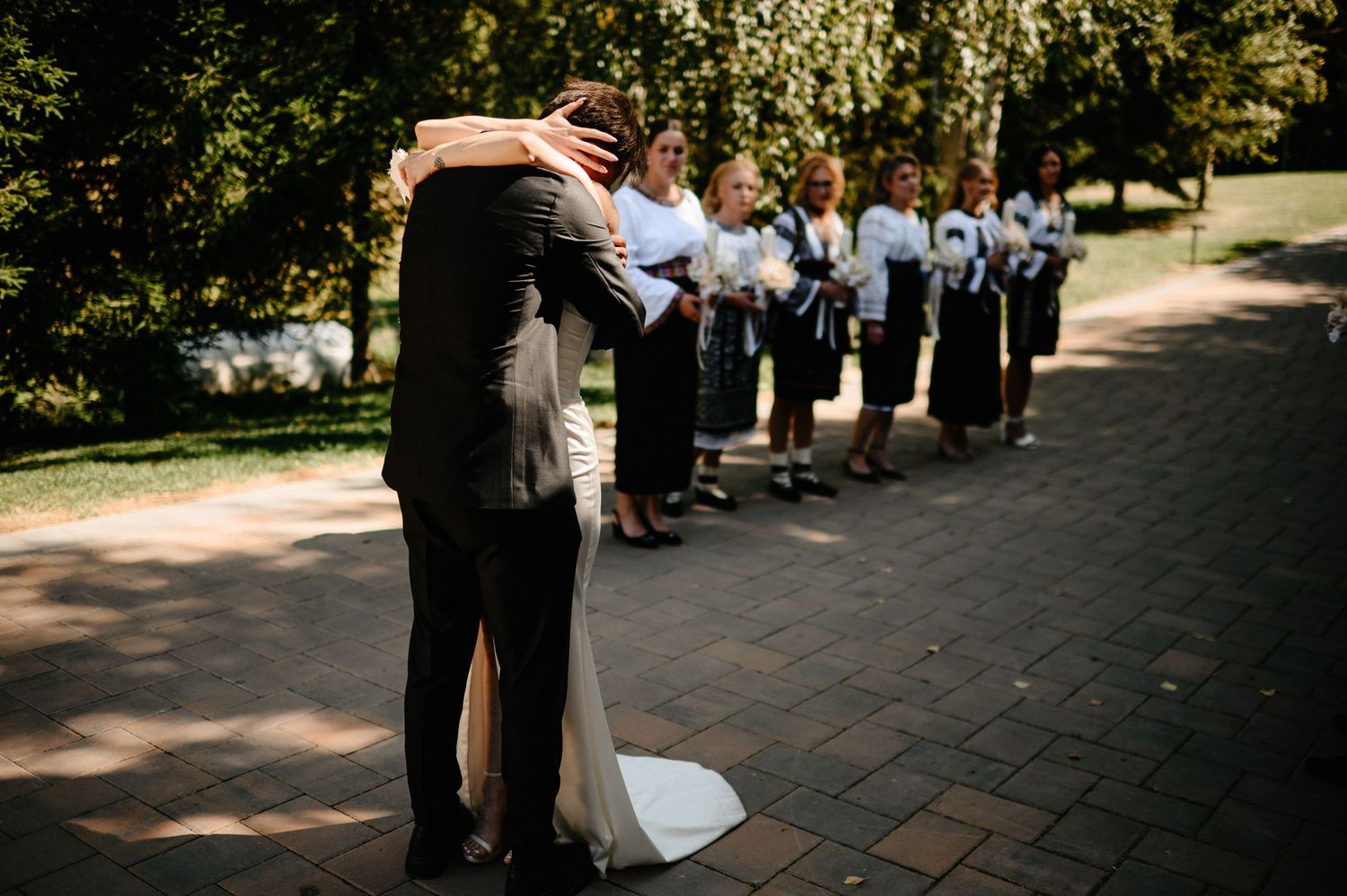 Bride and groom share first look hug on brick path with wedding party watching.