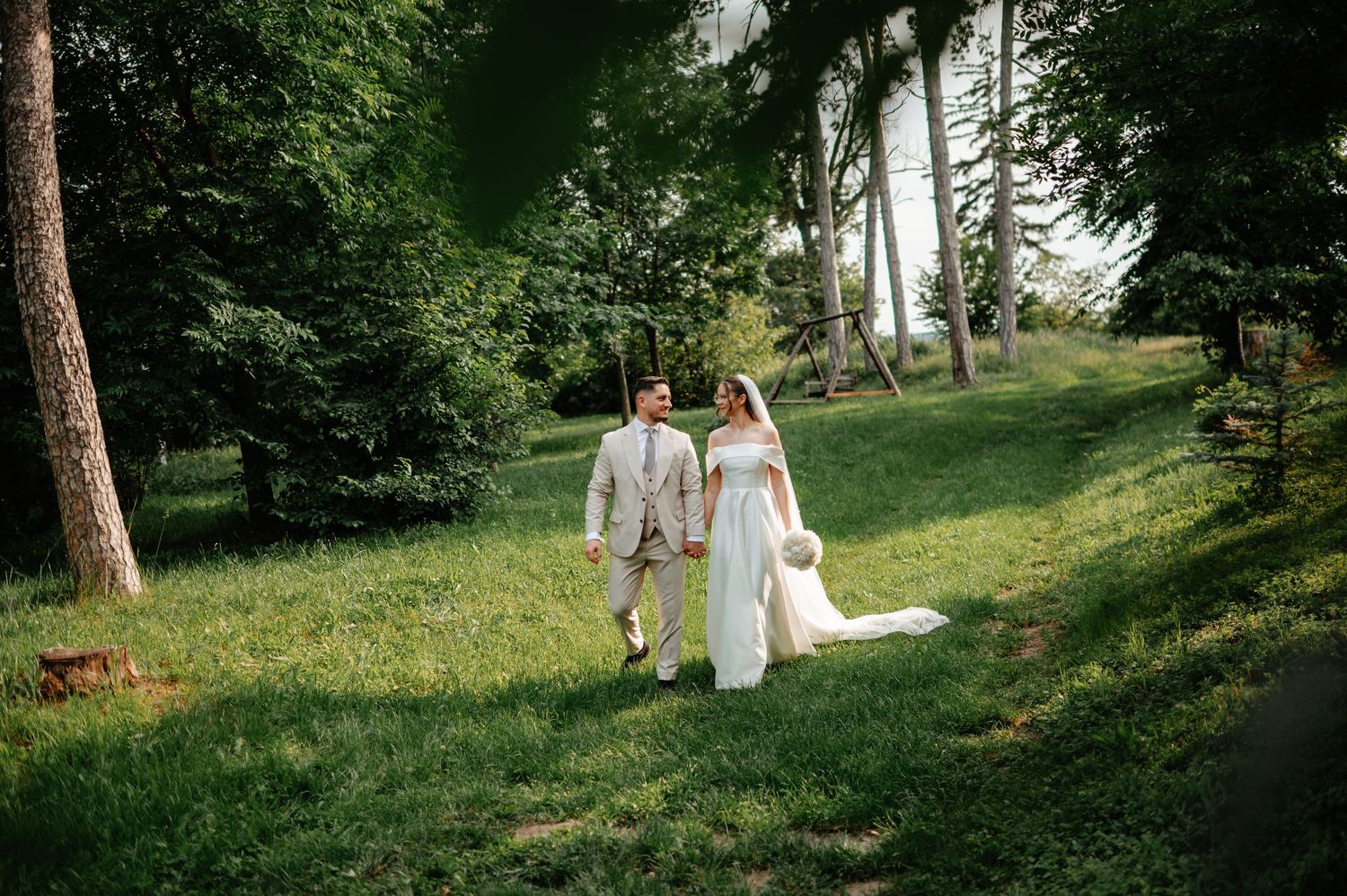 Bride and groom holding hands walking on lush green hillside surrounded by tall trees.