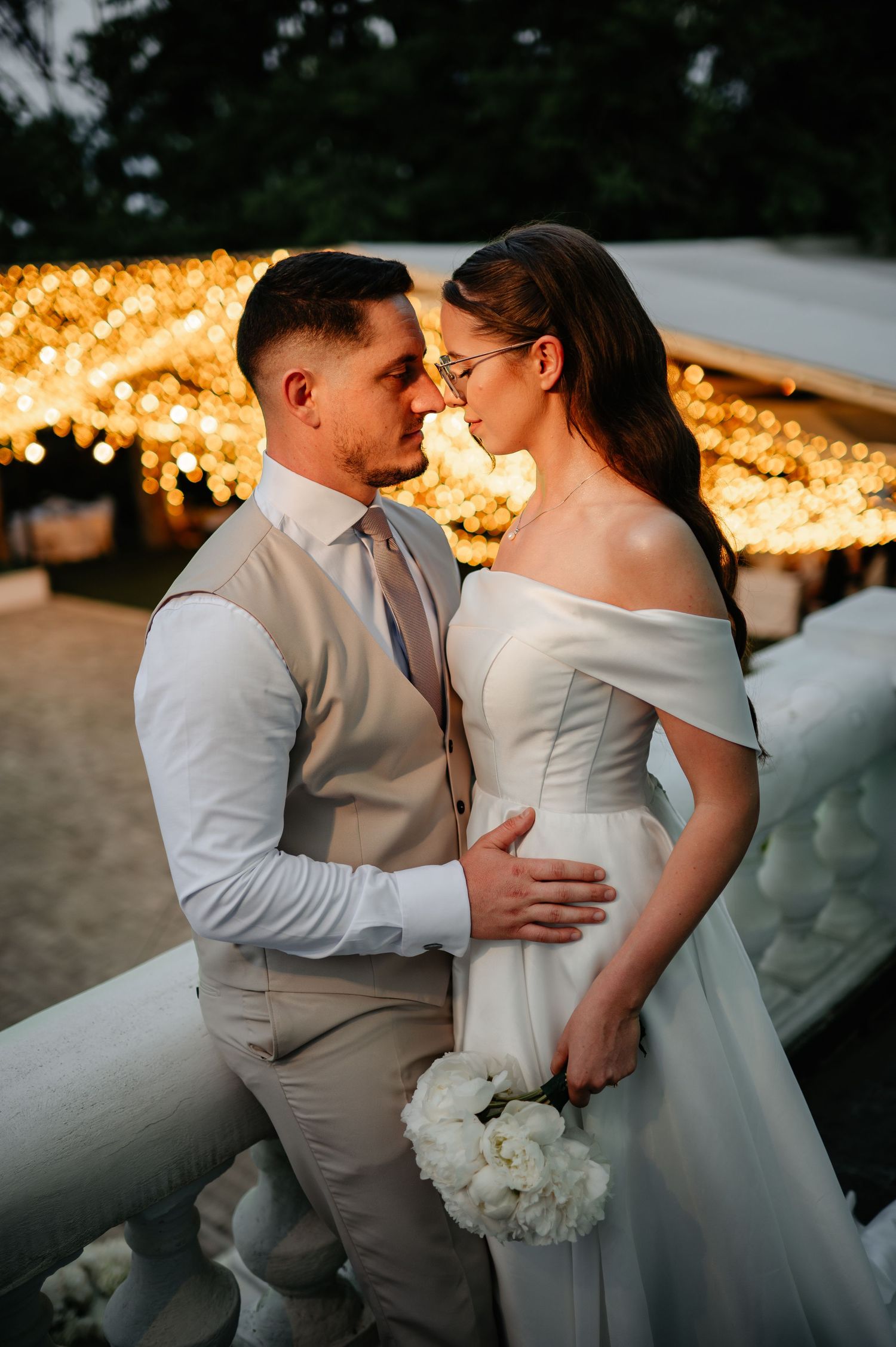 Bride and groom sharing a romantic moment in front of warm string lights at their outdoor wedding reception.