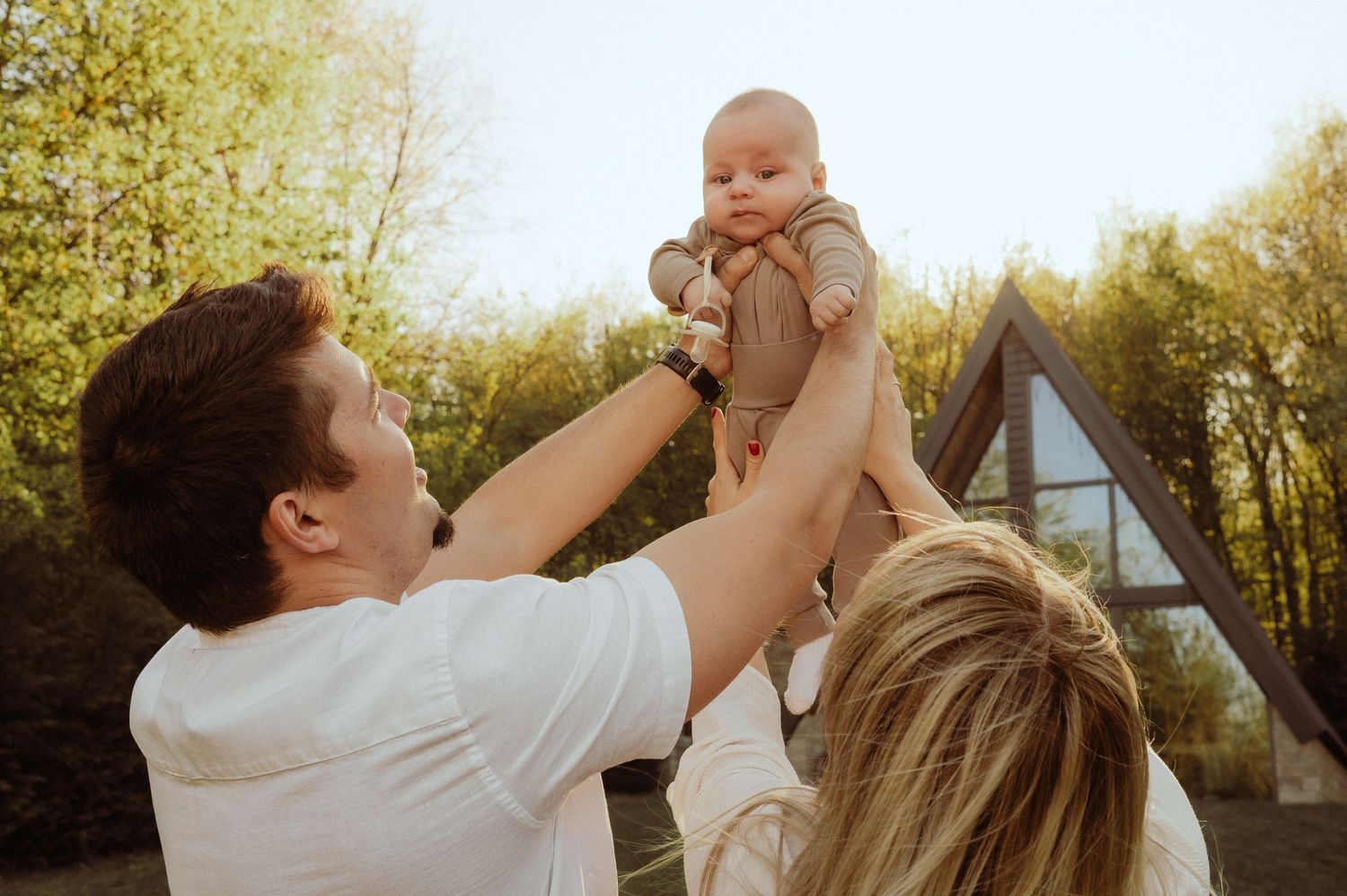 Father lifting smiling baby up in the air outdoors near an A-frame cabin in golden sunlight.
