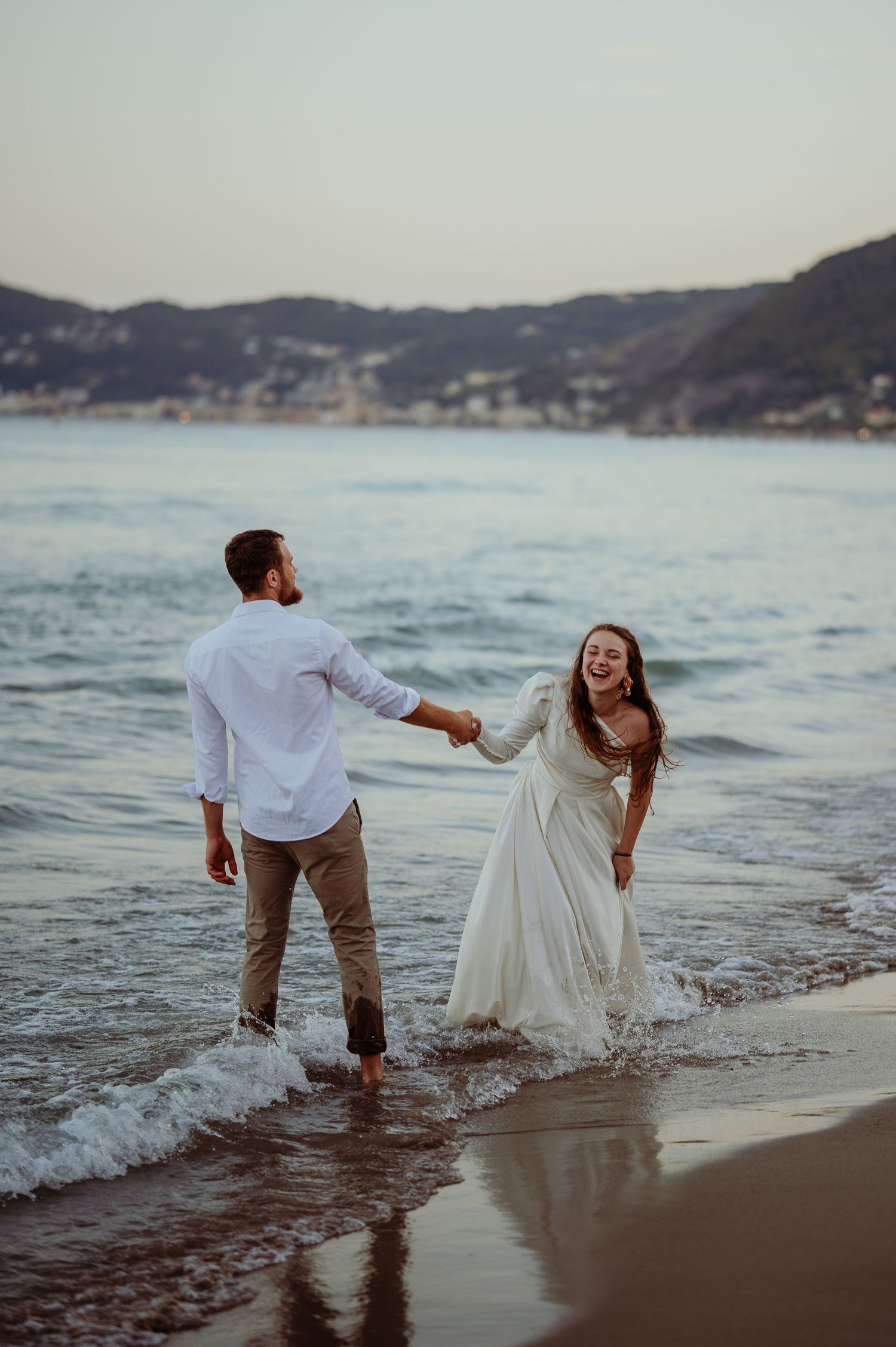 Laughing bride in white gown and groom walking through ocean waves on beach.