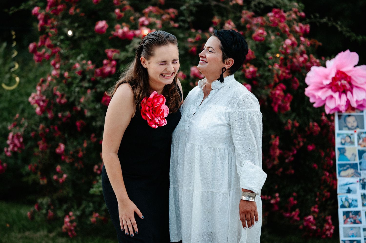 Two women laughing joyfully together in front of lush pink flowering rose bushes outdoors.