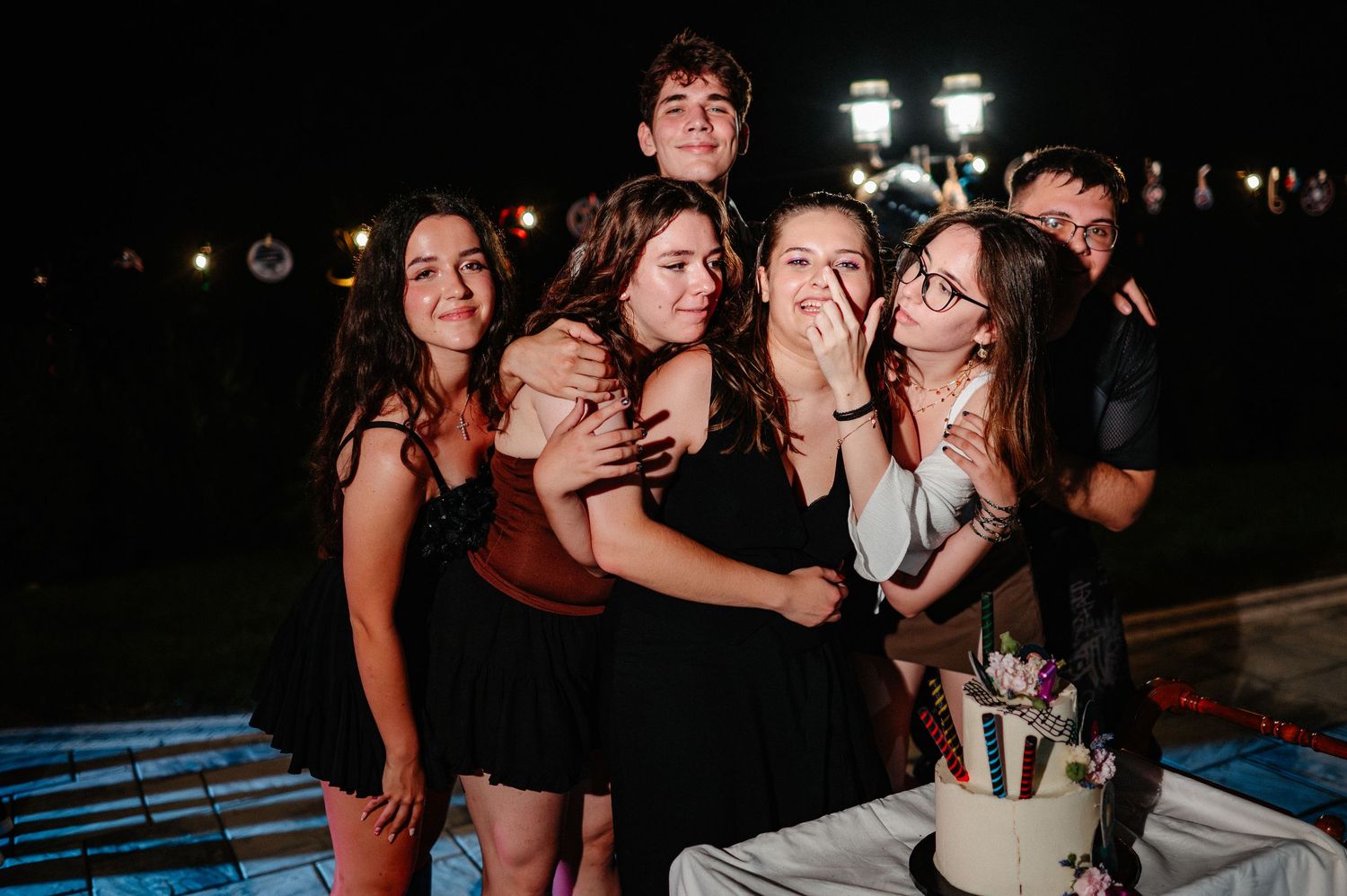 Group of six friends hugging and posing together at night celebration near a white birthday cake.