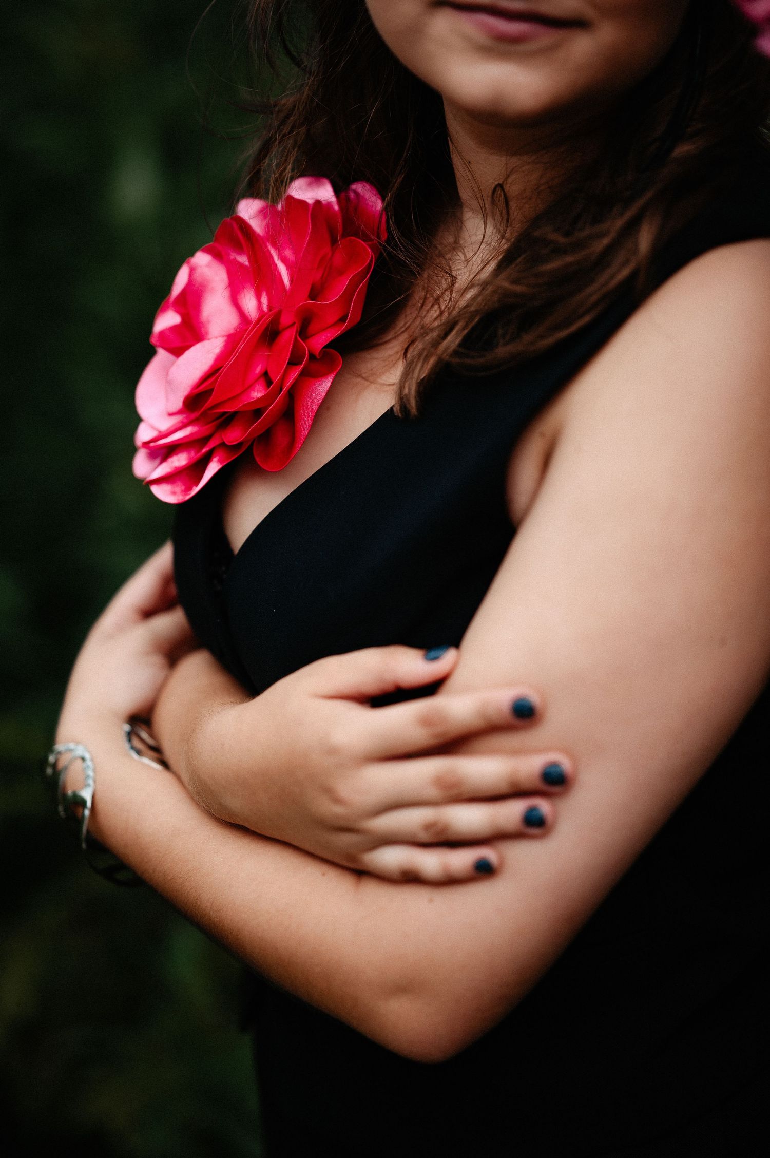 Young woman in black dress with red flower corsage crossing arms, dark nail polish, bracelet visible.
