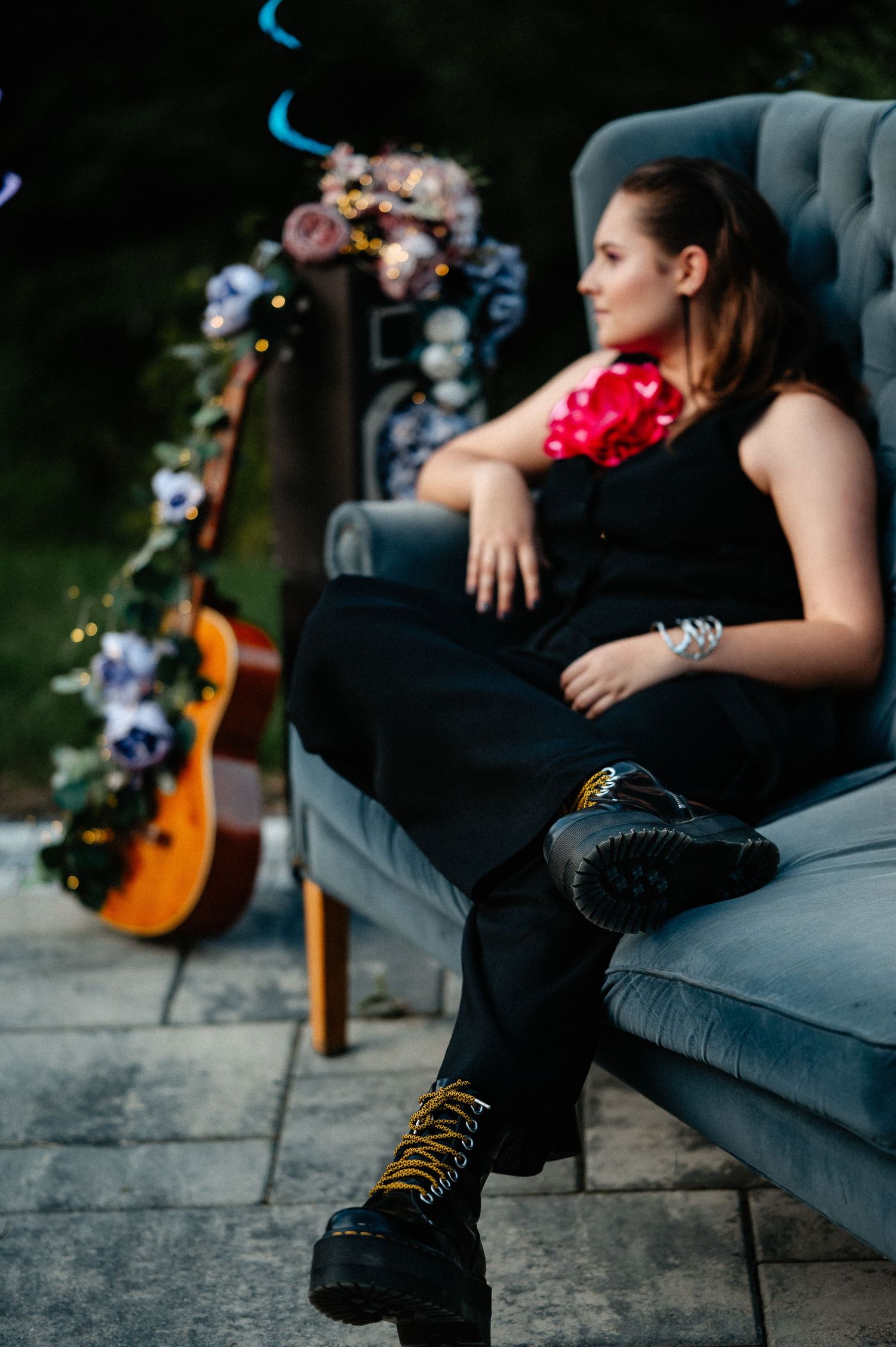 Young woman in black outfit with red corsage sitting in gray velvet chair beside a decorated guitar.