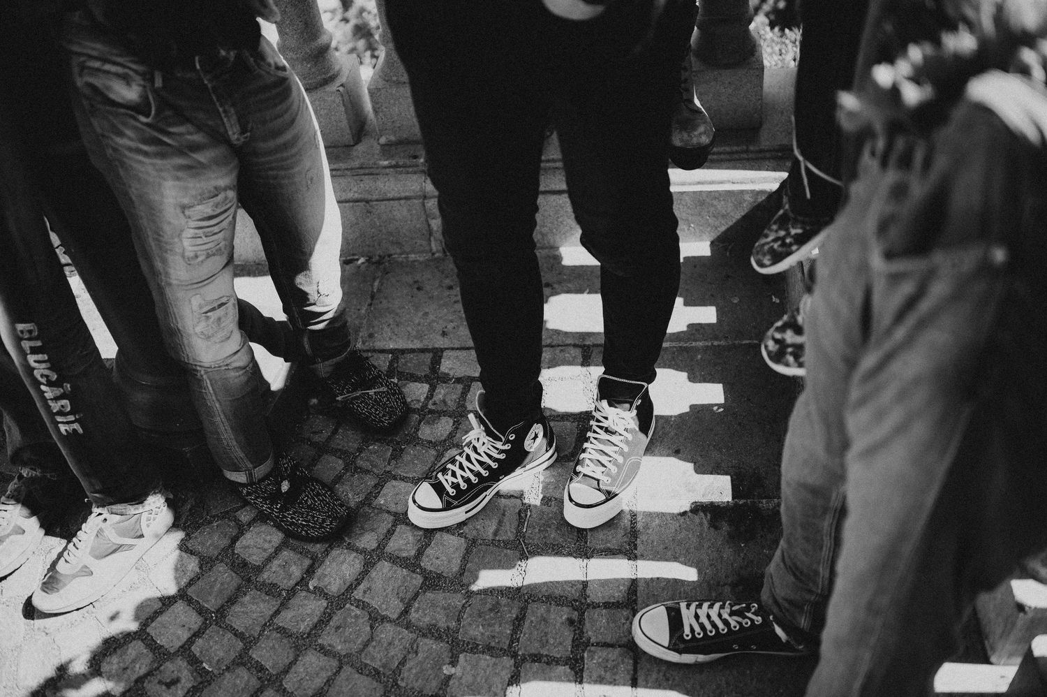 Black and white photo of group of people wearing Converse sneakers standing on cobblestone.