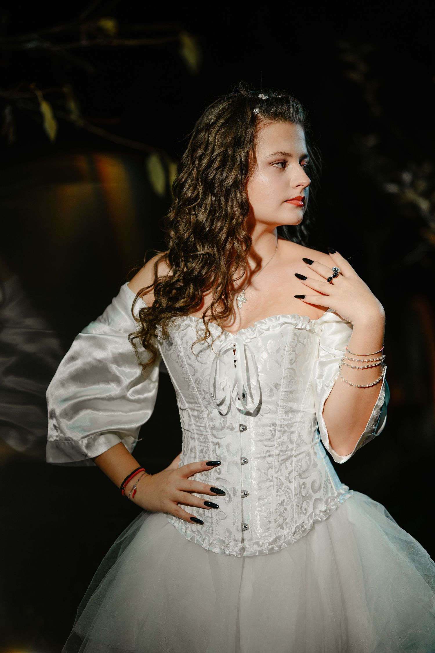 Young woman in white corset Halloween bride costume with black nails and curly hair posing outdoors at night.