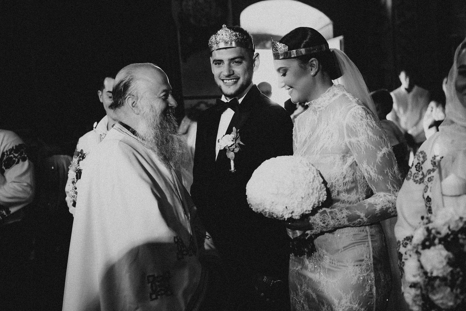Black and white photo of smiling couple with Orthodox bishop during traditional wedding ceremony.